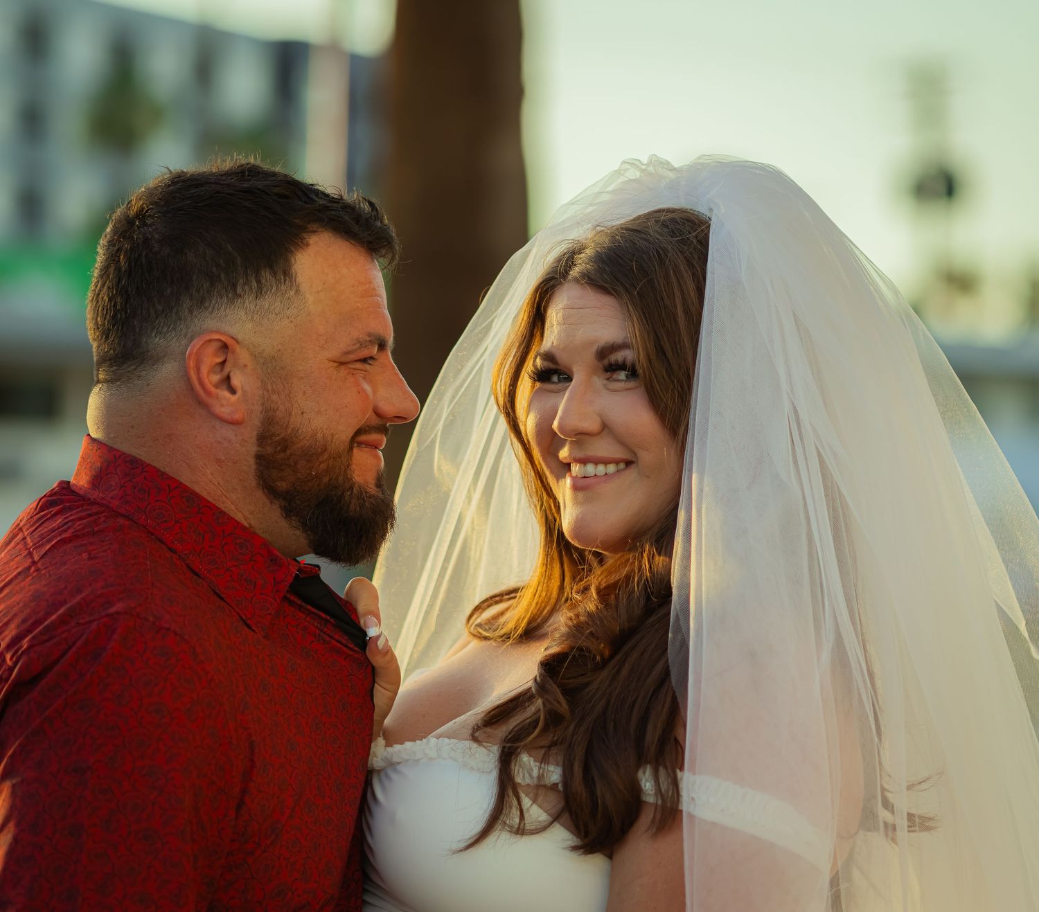 Bride and groom sharing a tender moment outdoors, groom in red shirt smiling at bride.