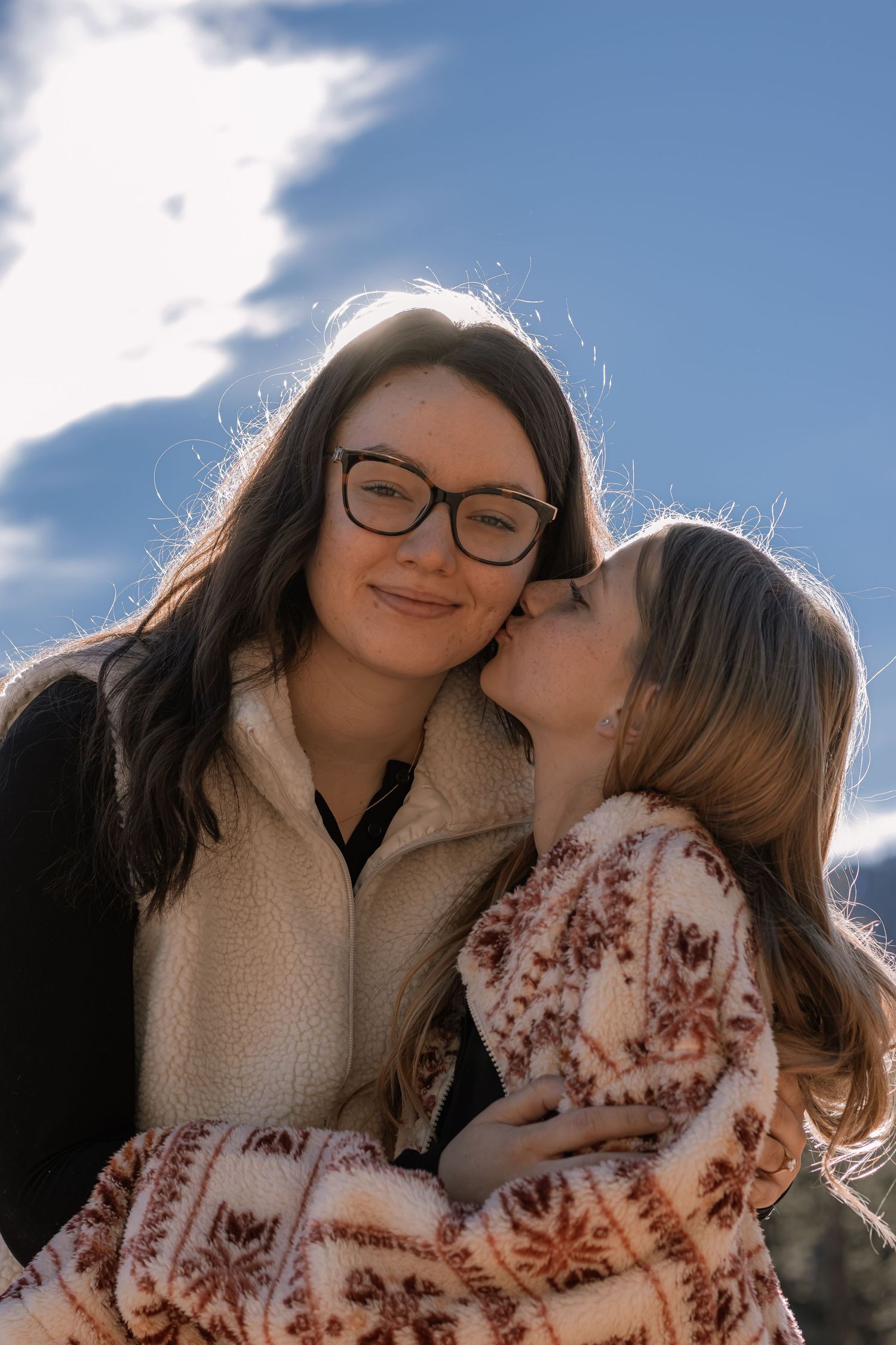 A woman with glasses smiles as a young girl kisses her cheek against a blue sky.
