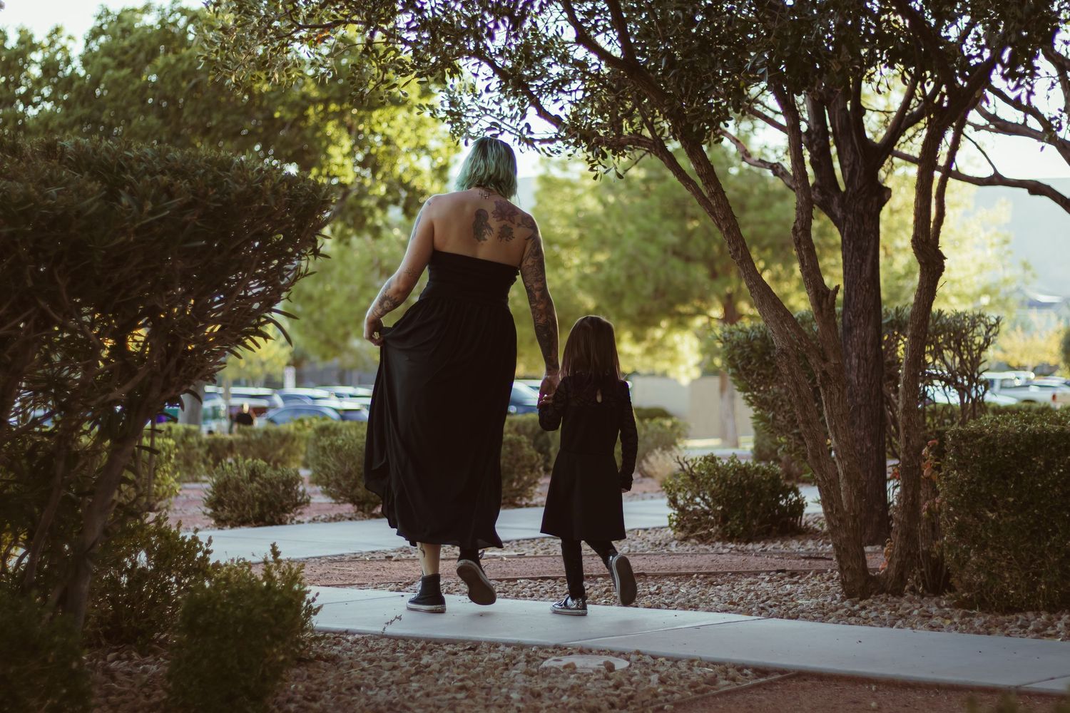 A woman with teal hair in a black dress walks hand-in-hand with a young child along a sunny sidewalk.