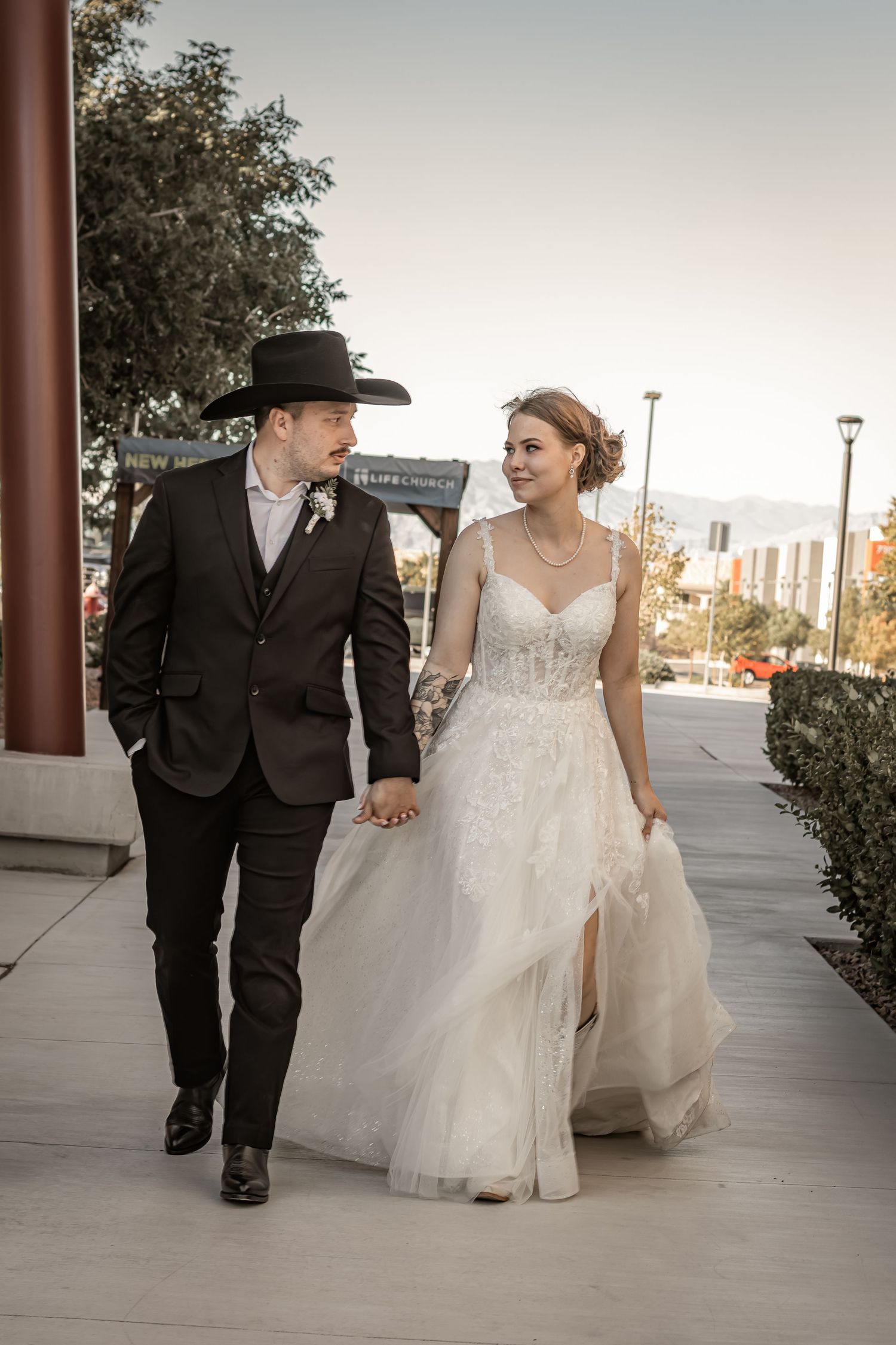 Bride and groom walk hand in hand, groom wearing black cowboy hat and suit, bride in lace wedding gown.