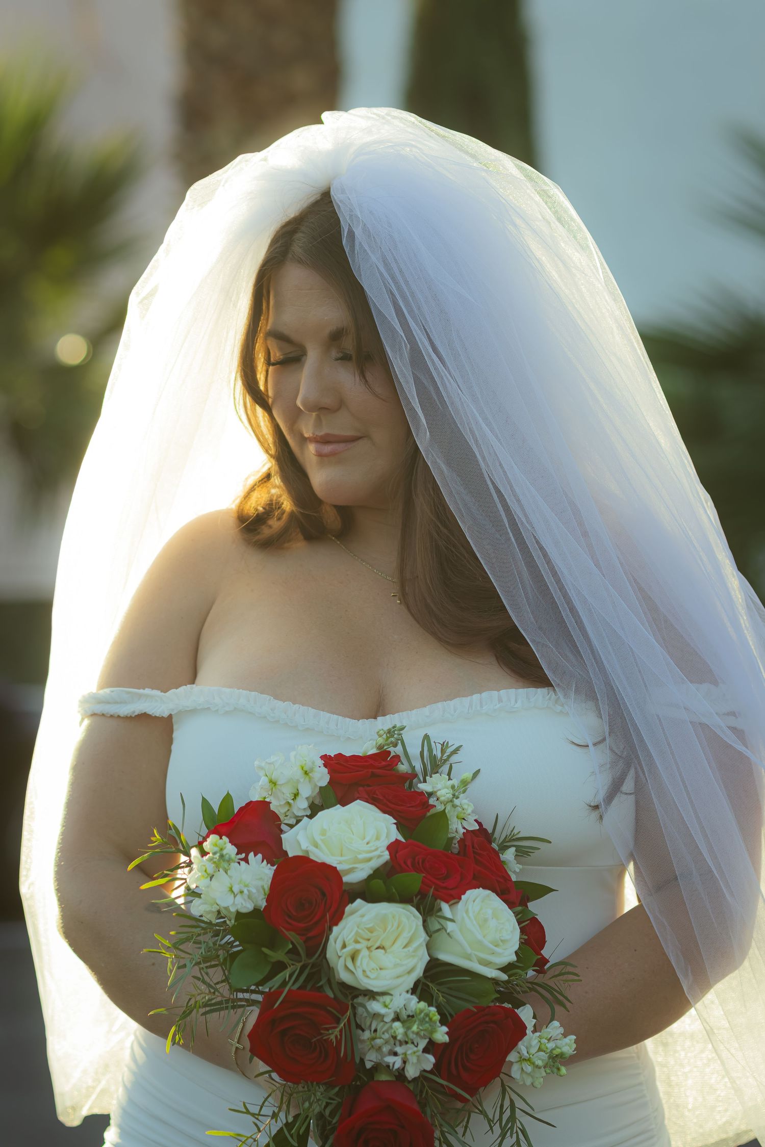 Bride in white off-shoulder gown holding red and white rose bouquet under flowing veil.