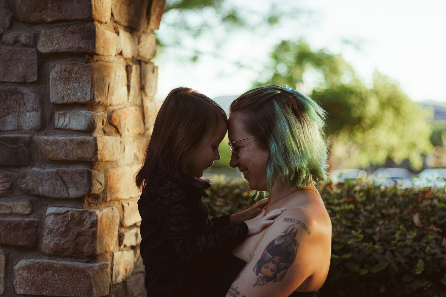 A tattooed woman with teal hair and a young girl touch foreheads tenderly beside a stone pillar outdoors.