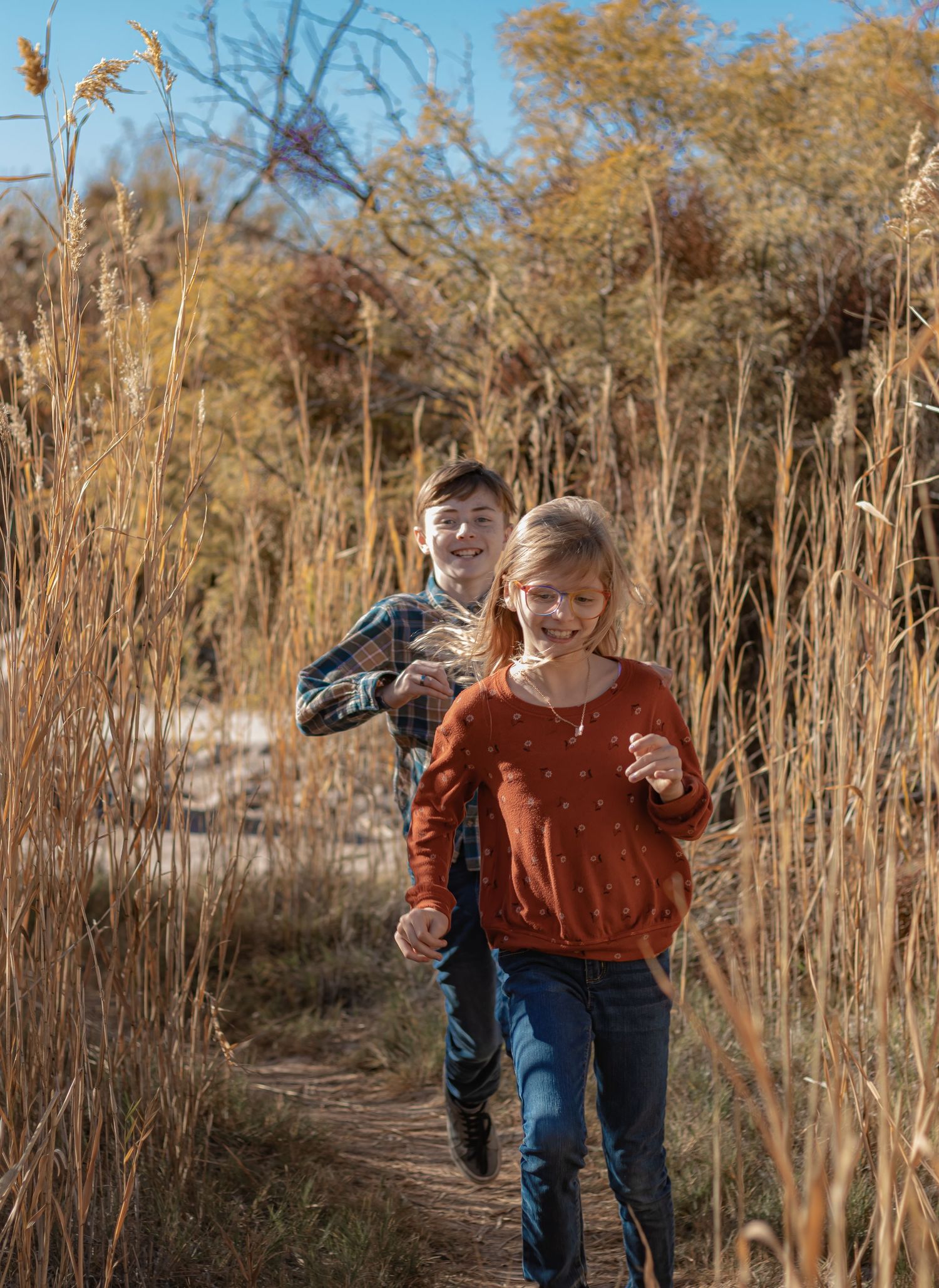 Two smiling children run joyfully along a narrow path through tall golden grass on a bright autumn day.