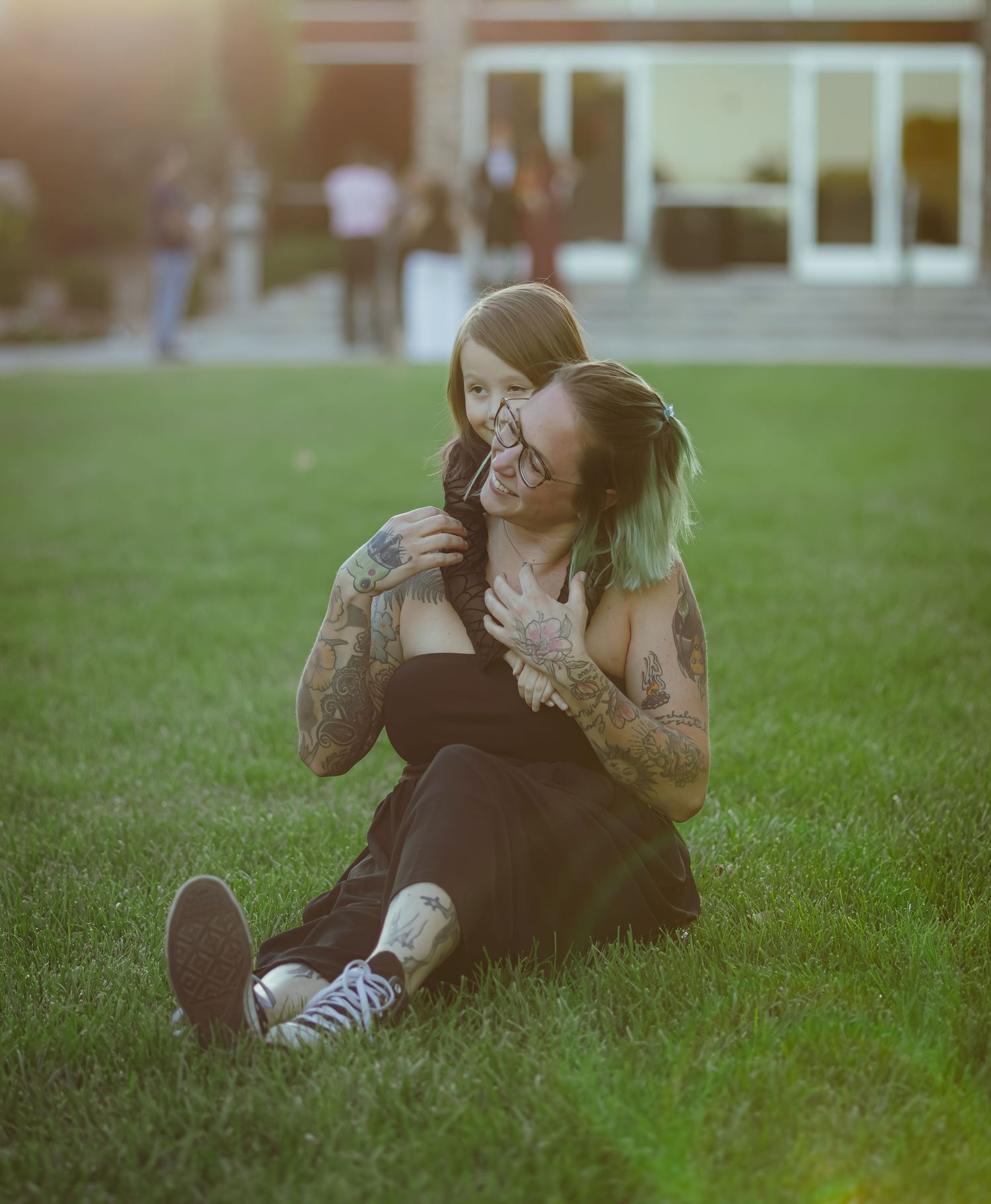 A tattooed woman and young child sit together on a grassy lawn, laughing and embracing in warm sunlight.