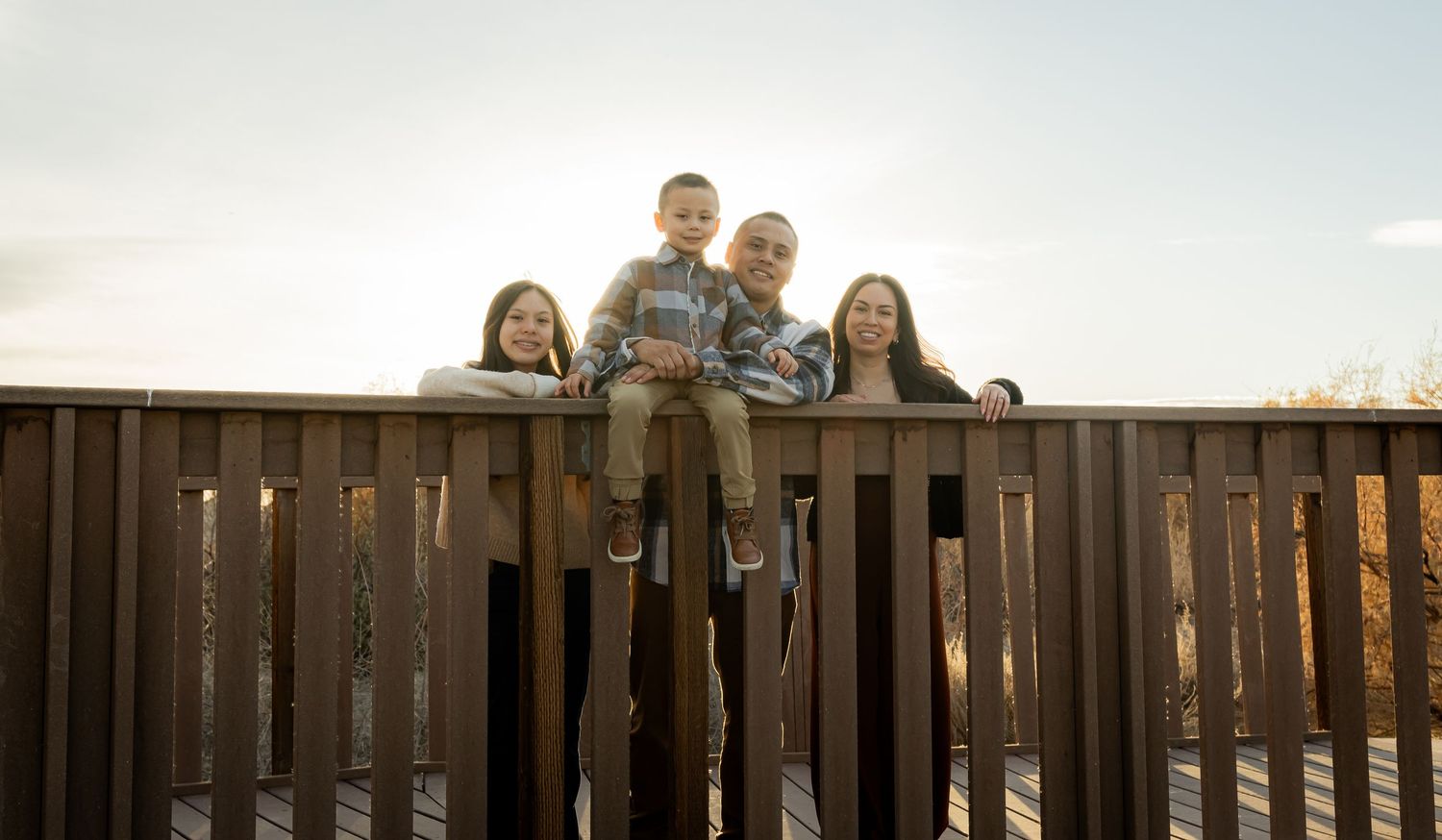 Family of four posing on a wooden deck railing with a bright sky and autumn foliage in the background.