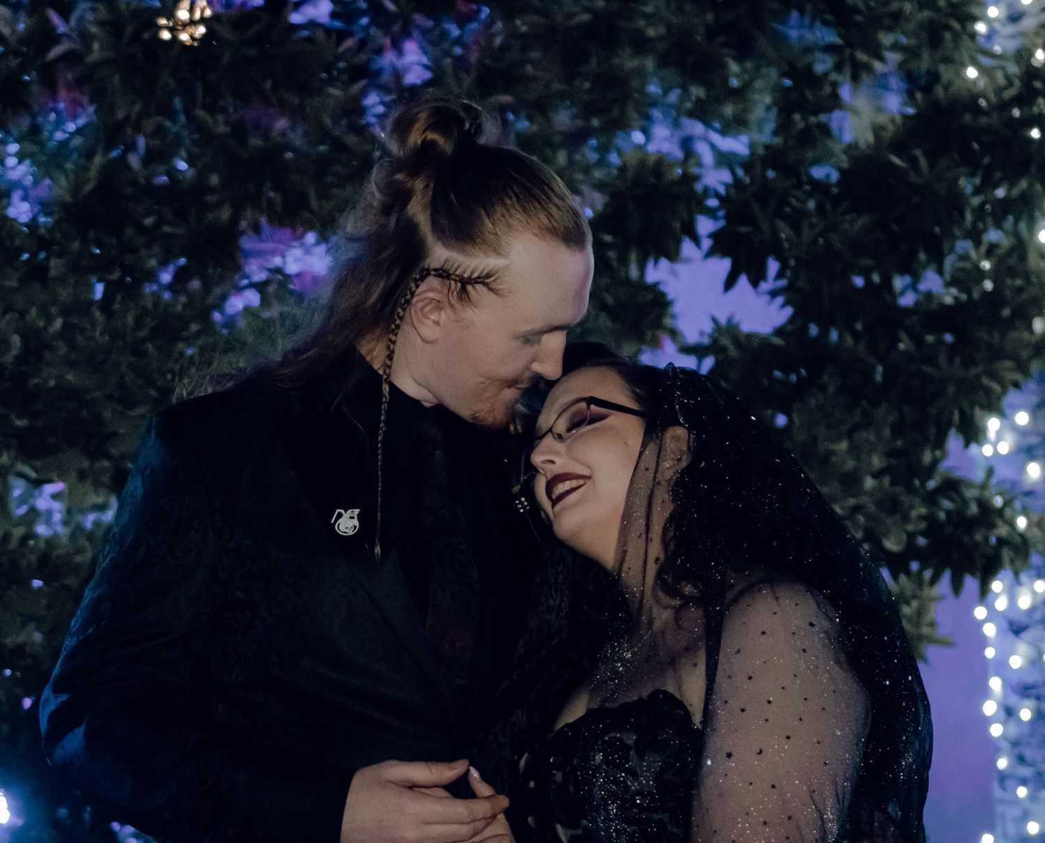 Gothic wedding couple gazes lovingly at each other under twinkling lights at night ceremony.