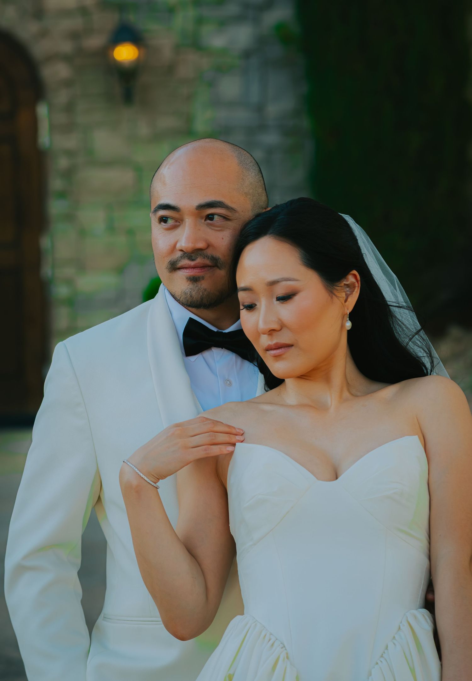 Asian wedding couple in classic white tuxedo and strapless gown posing elegantly before stone wall at dusk.