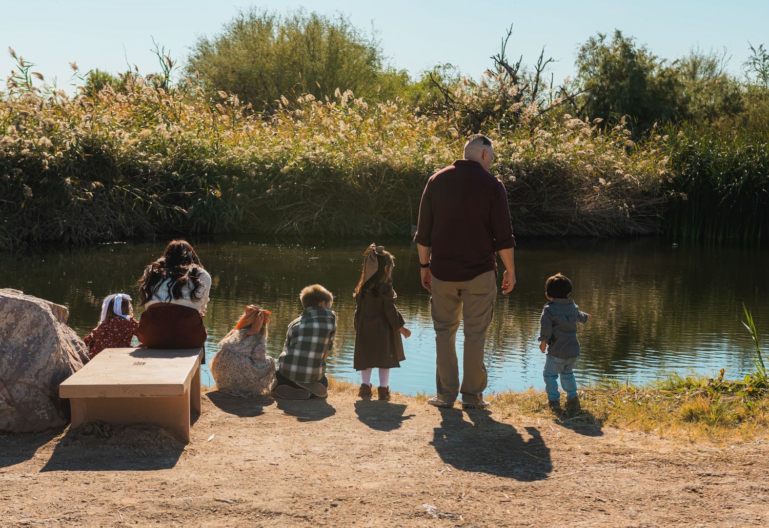 A family gathers along the edge of a calm pond, with children sitting and a man walking near the water.