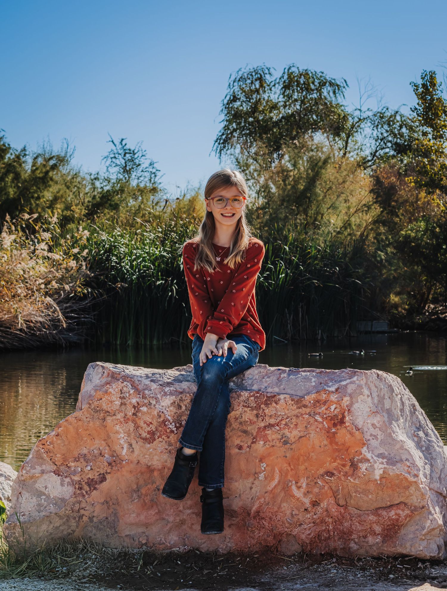 A teenage girl in a red top and ripped jeans sits on a large rock near a pond.