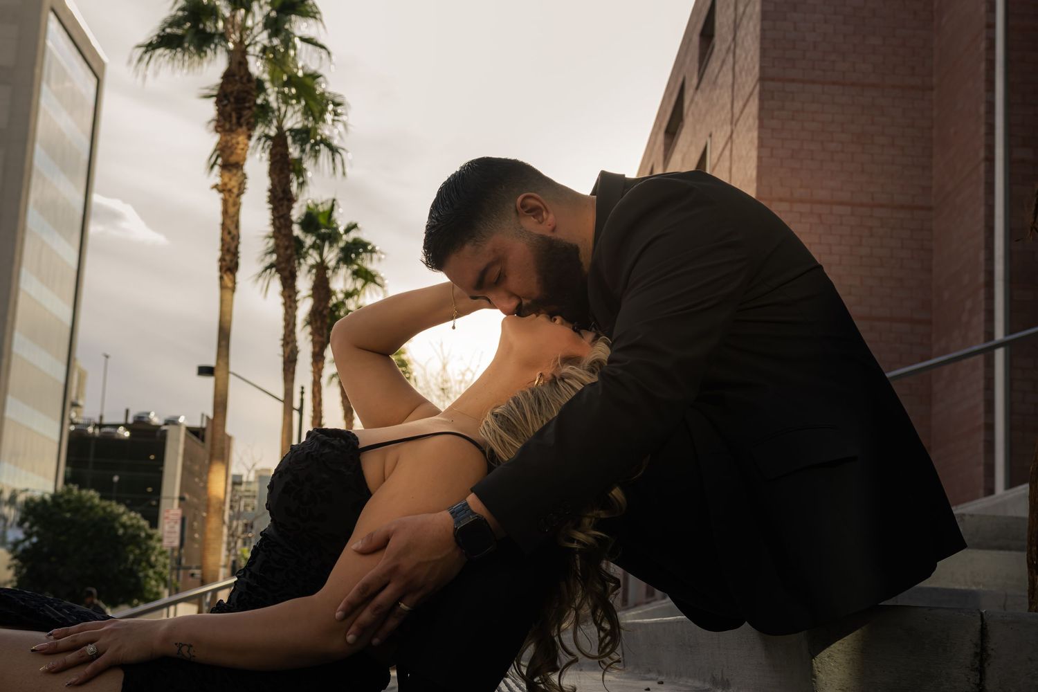 Intimate moment of a bearded man embracing a tattooed woman on steps near palm trees.