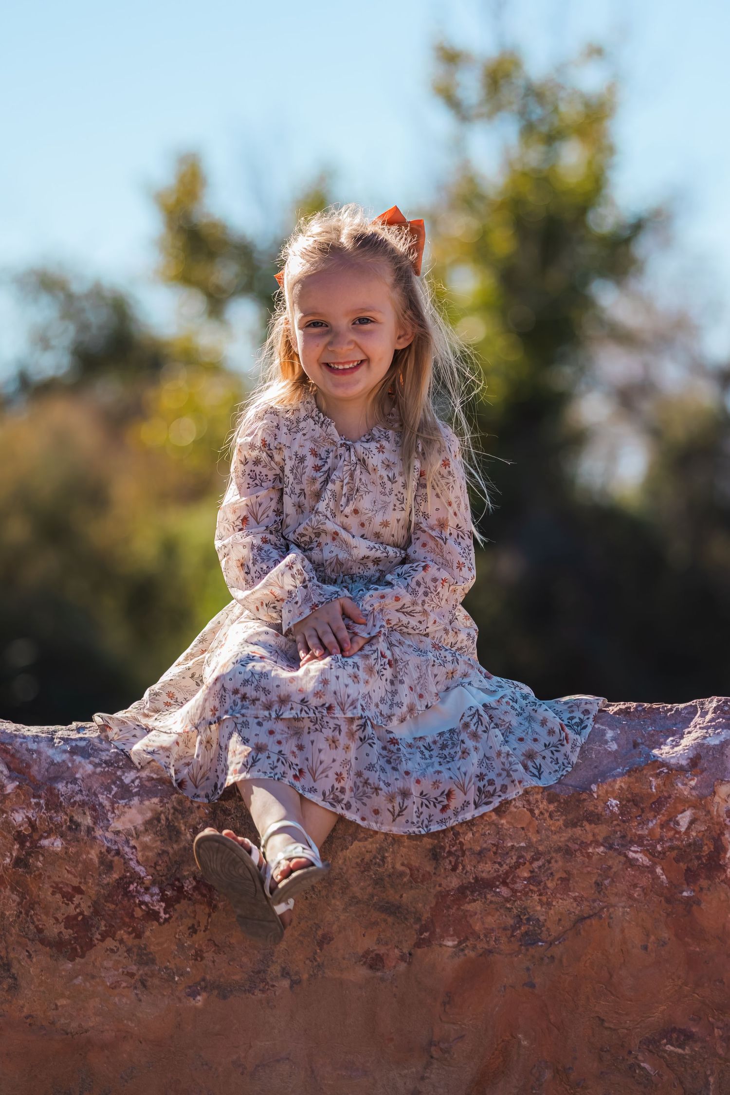 Smiling blonde girl in a floral dress sitting on a rock outdoors.