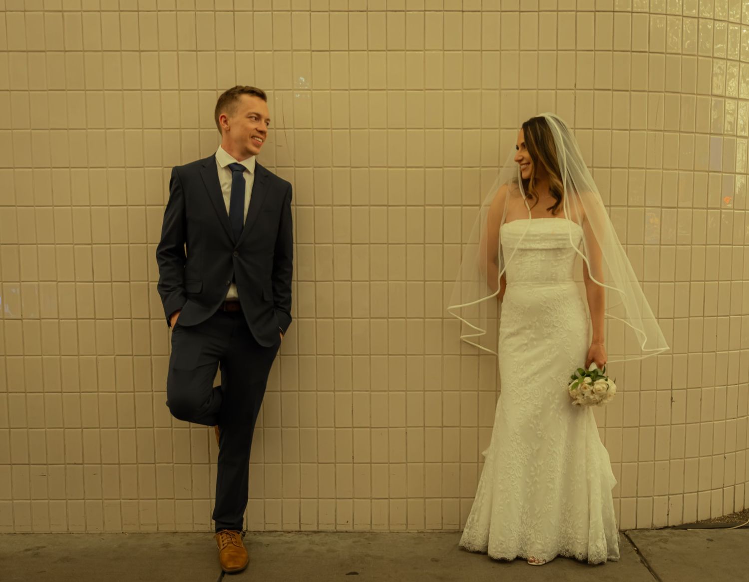 Bride in white gown and groom in navy suit share a joyful moment against a tiled wall.