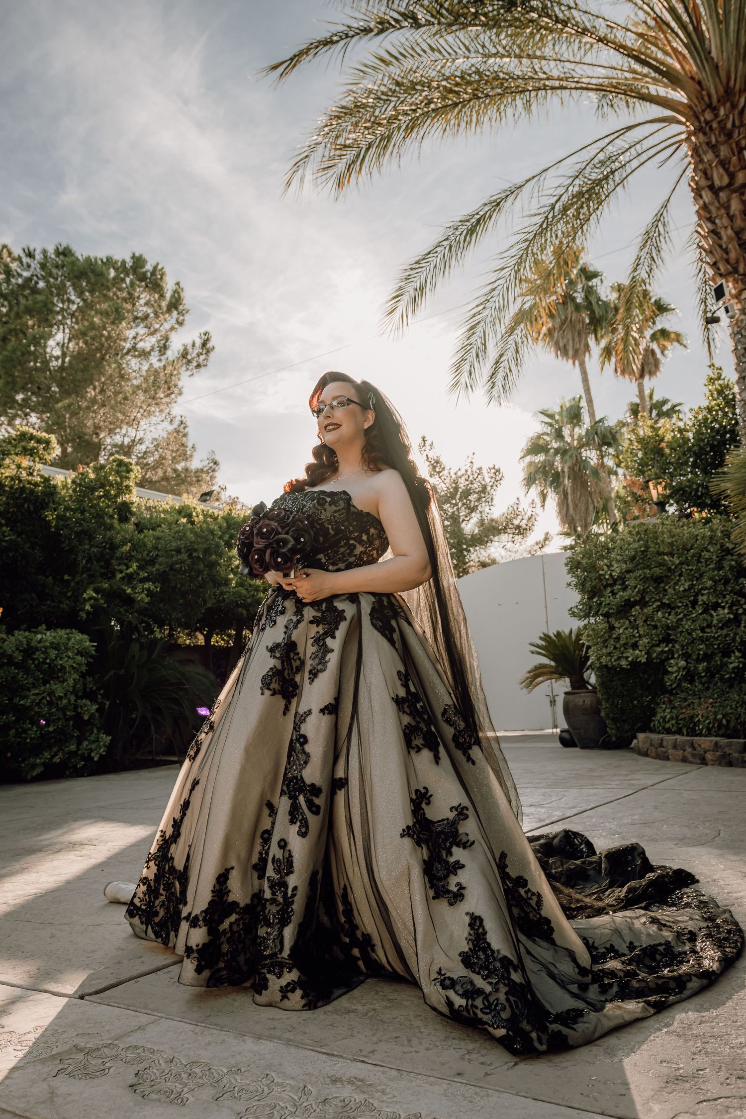Bride in stunning black lace applique champagne ballgown holding dark bouquet under palm trees.