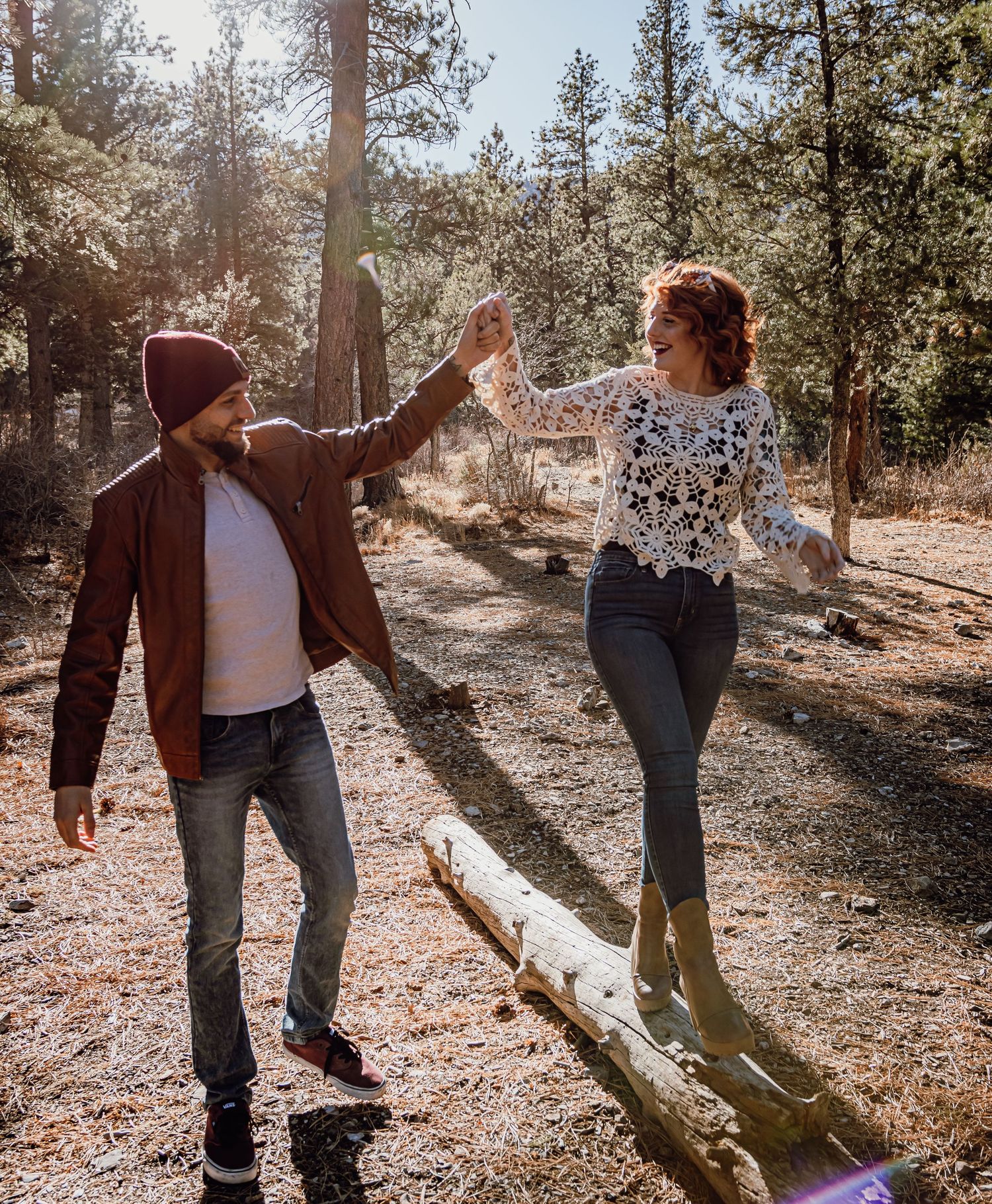 A couple dances joyfully on a fallen log in a sunny pine forest, the man holding the woman's hand as she balances.