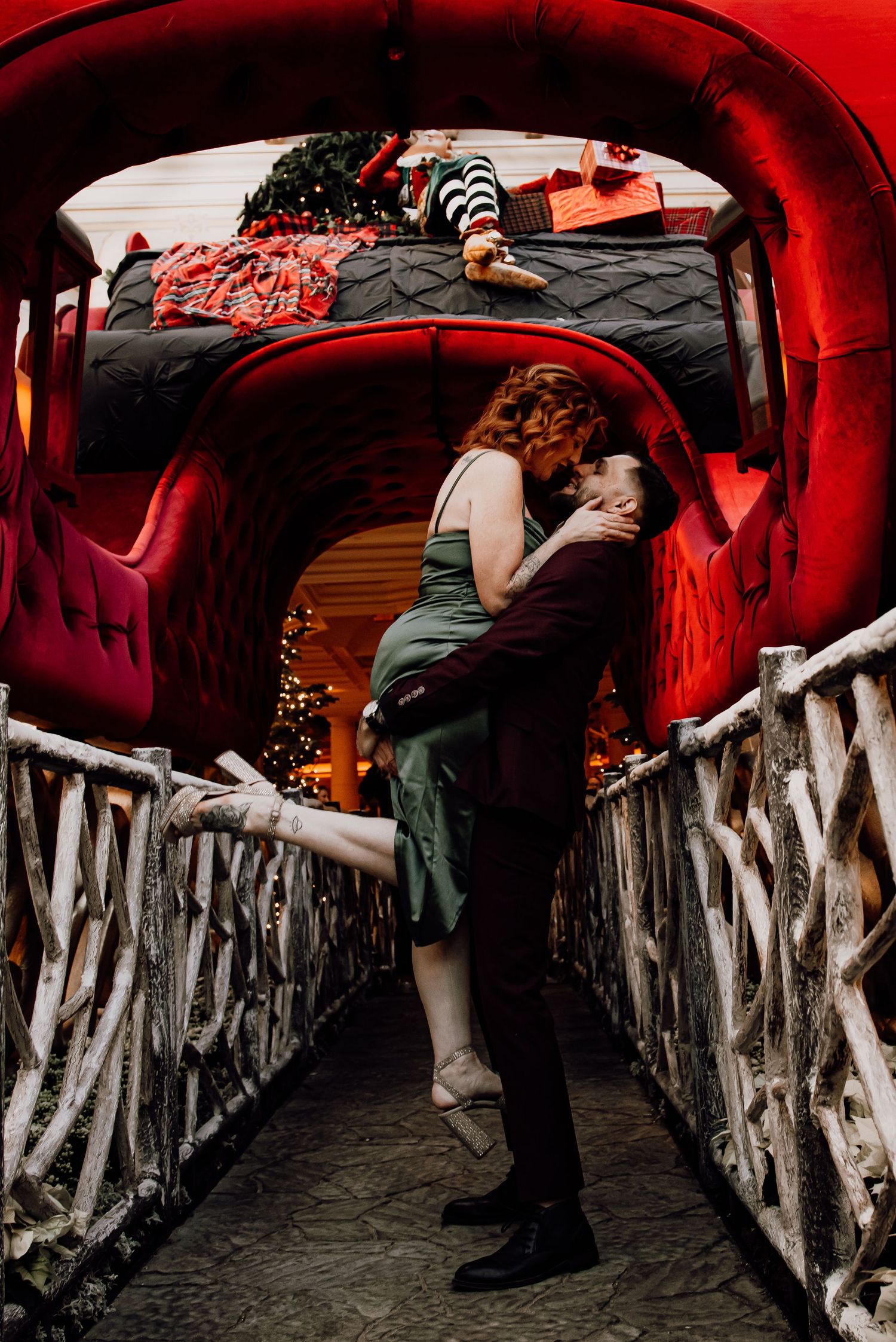 Couple sharing a romantic kiss on a festive bridge with red sleigh and Christmas decorations in background.