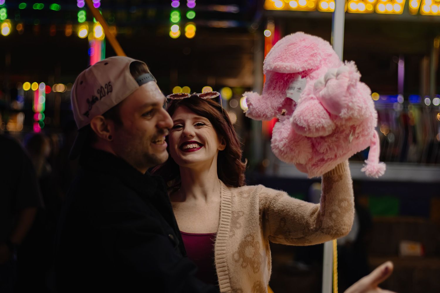 Couple smiling at a carnival at night, holding a large pink stuffed animal prize under colorful lights.