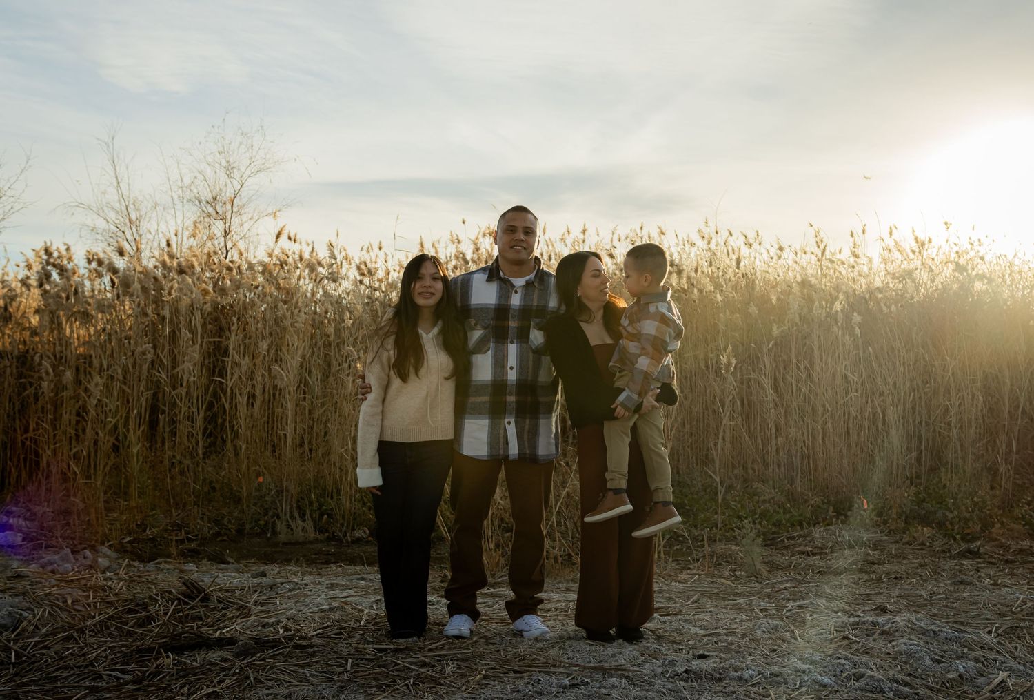 Family of four smiling together in front of tall golden reeds during a warm sunset outdoor portrait session.