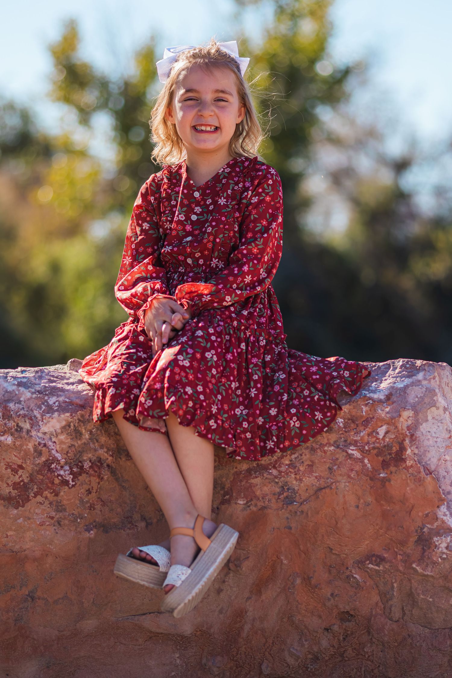 Smiling girl in a red floral dress sitting on a rock outdoors.