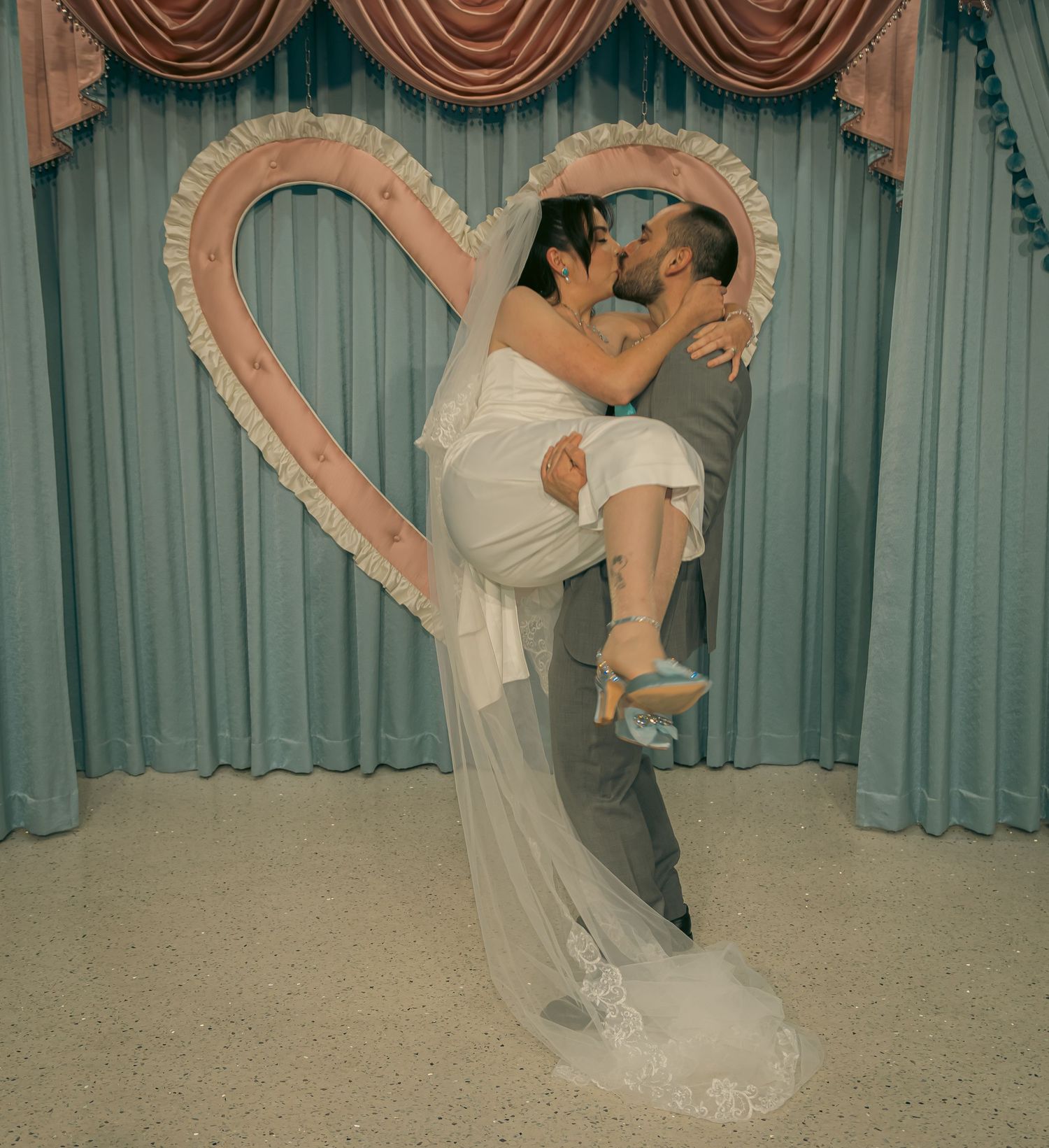 Groom lifts bride for a passionate kiss in front of a decorative heart backdrop at a wedding chapel.