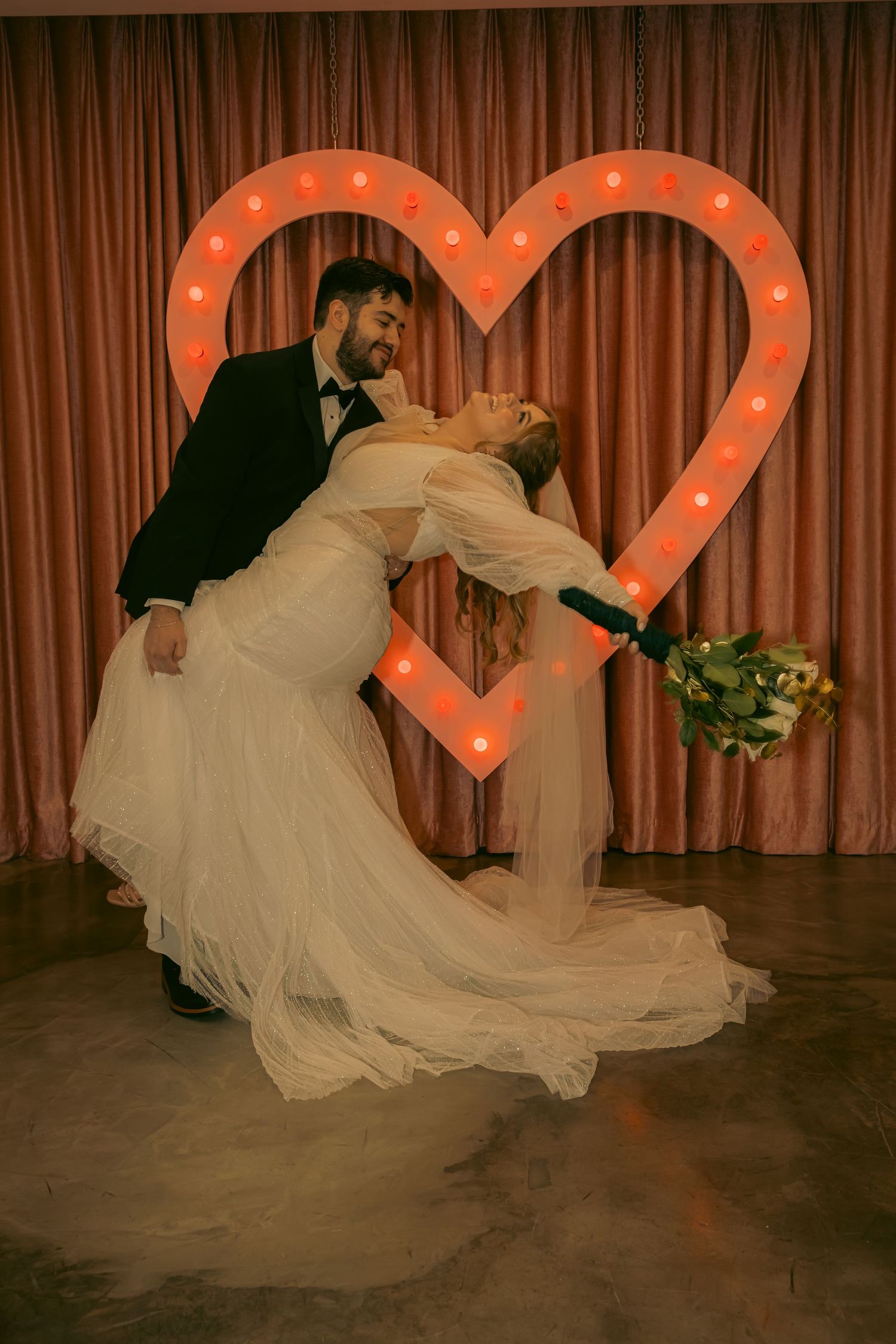 Newlyweds share a romantic dip kiss in front of a glowing marquee heart backdrop, bride holding a bouquet.
