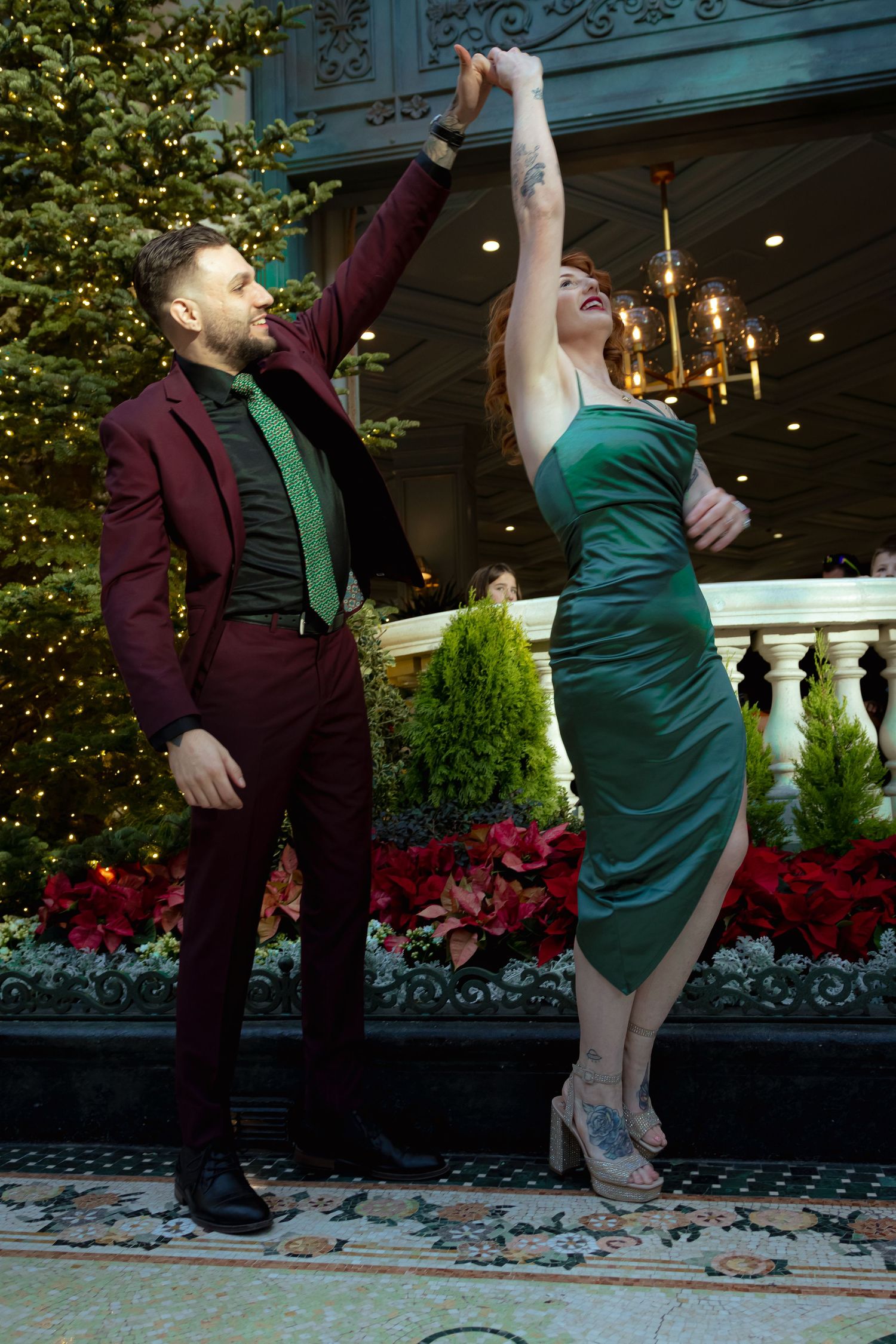 Couple dancing elegantly in festive holiday attire, man in burgundy suit, woman in green dress, near Christmas trees.