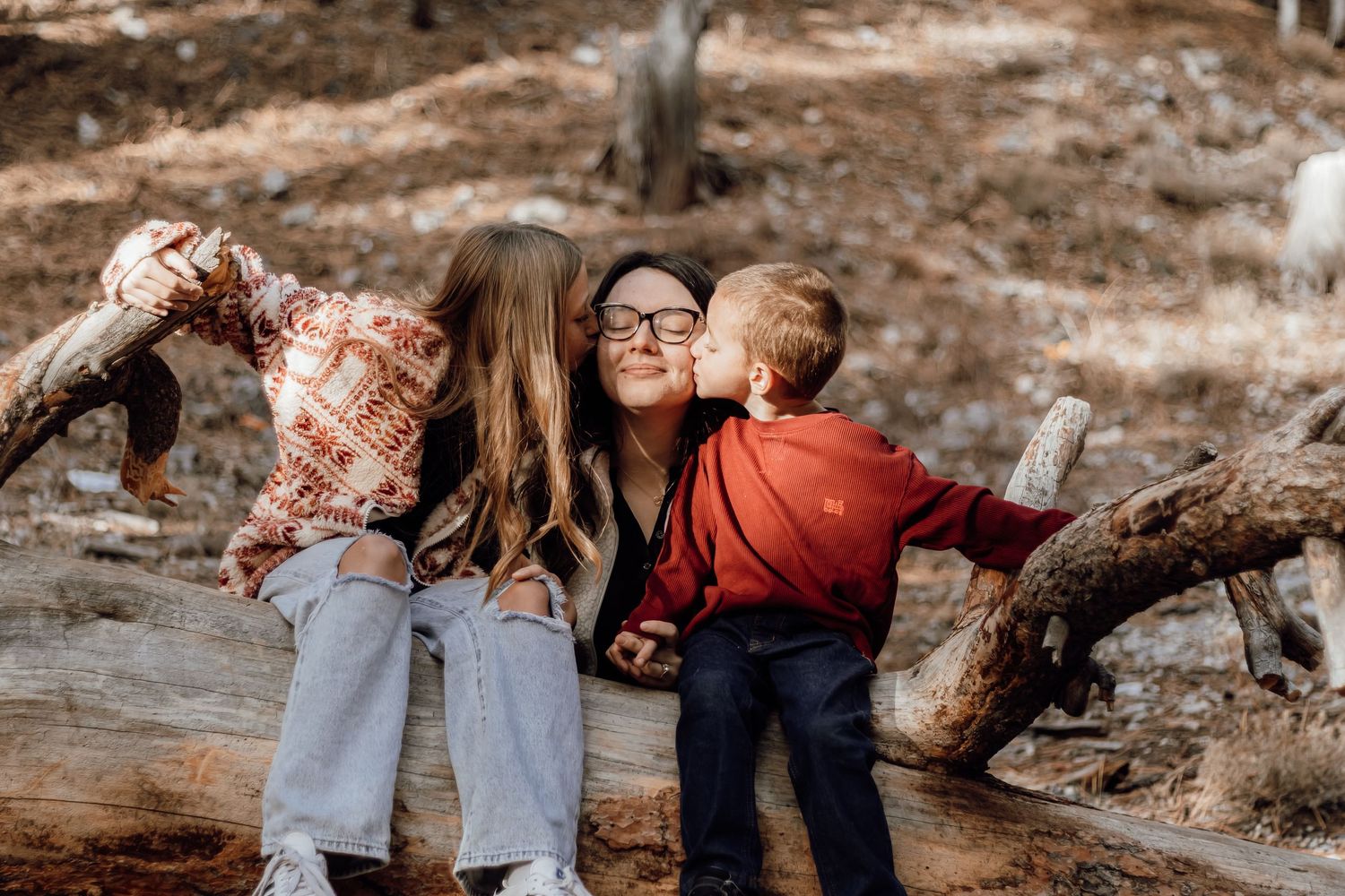 Mother being kissed on both cheeks by her two children while sitting on a log outdoors.