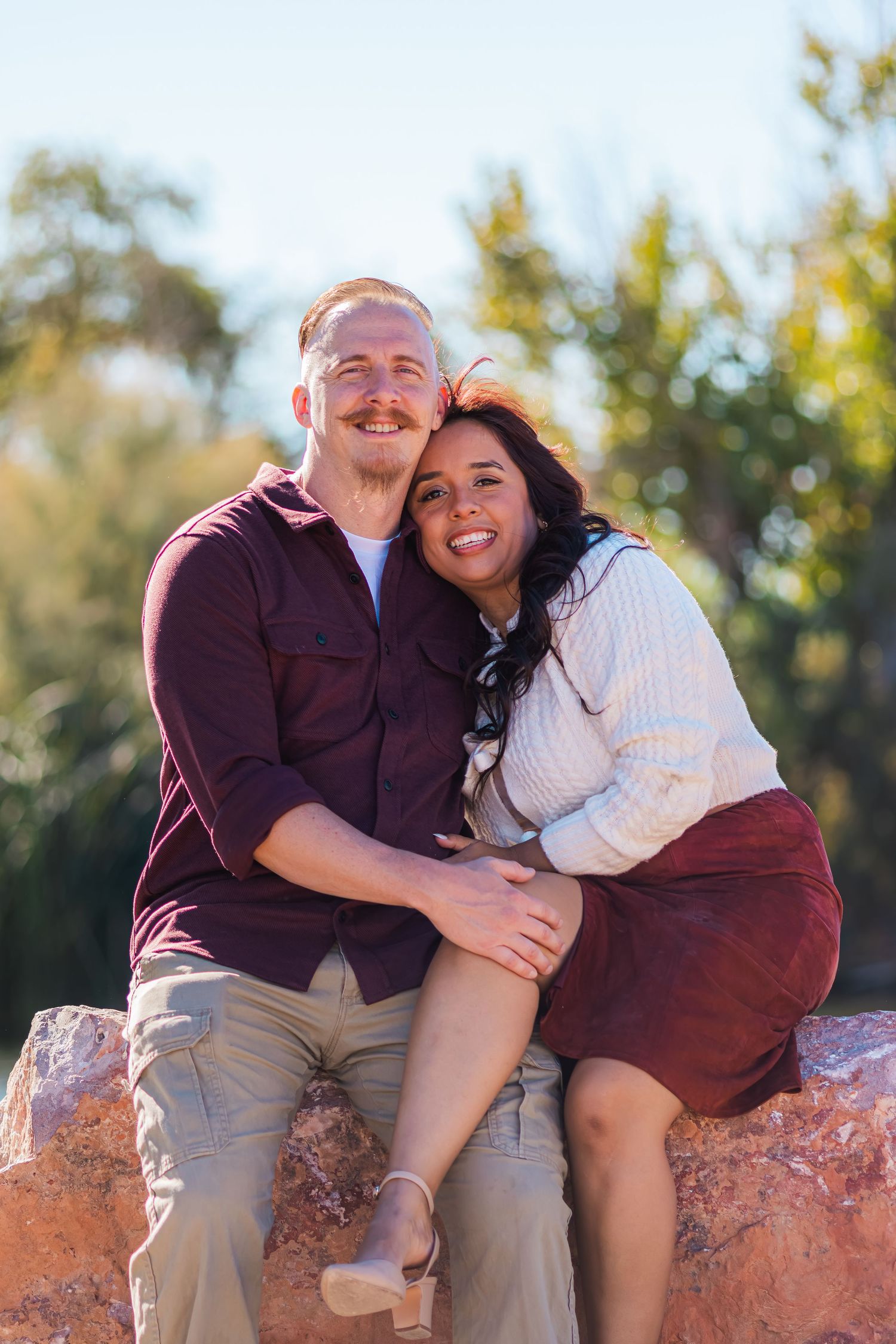 Smiling couple sitting closely together on rocks in a sunny outdoor setting.