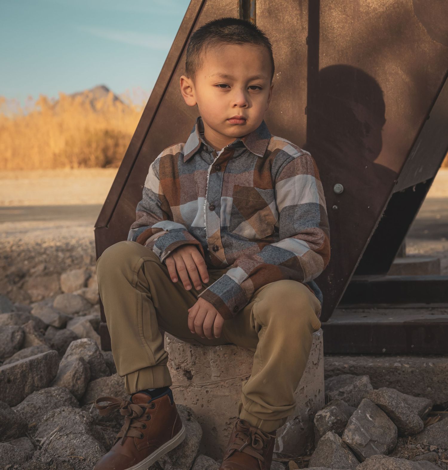 Young boy in a plaid shirt and khaki pants sitting on rocks near a metal structure in a desert setting.