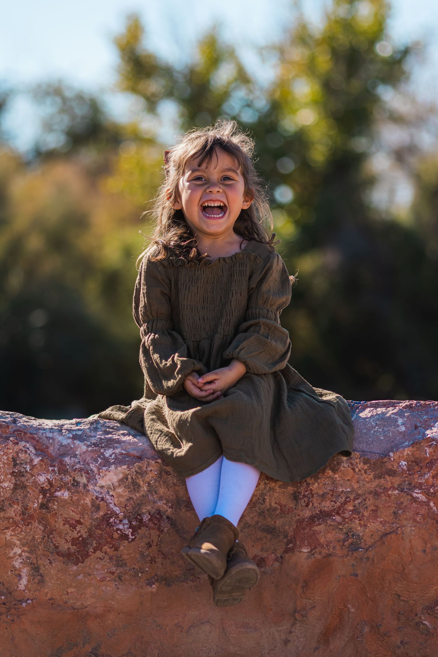 Laughing young girl in an olive dress sitting on a rock outdoors.