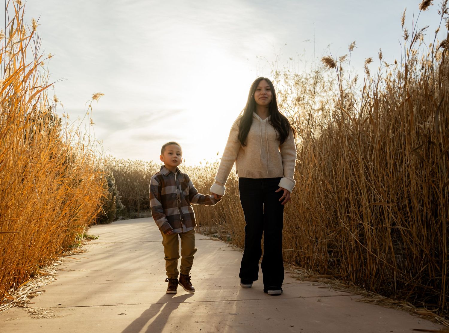 Young woman and little boy walking hand in hand on a sunny path lined with golden autumn reeds.
