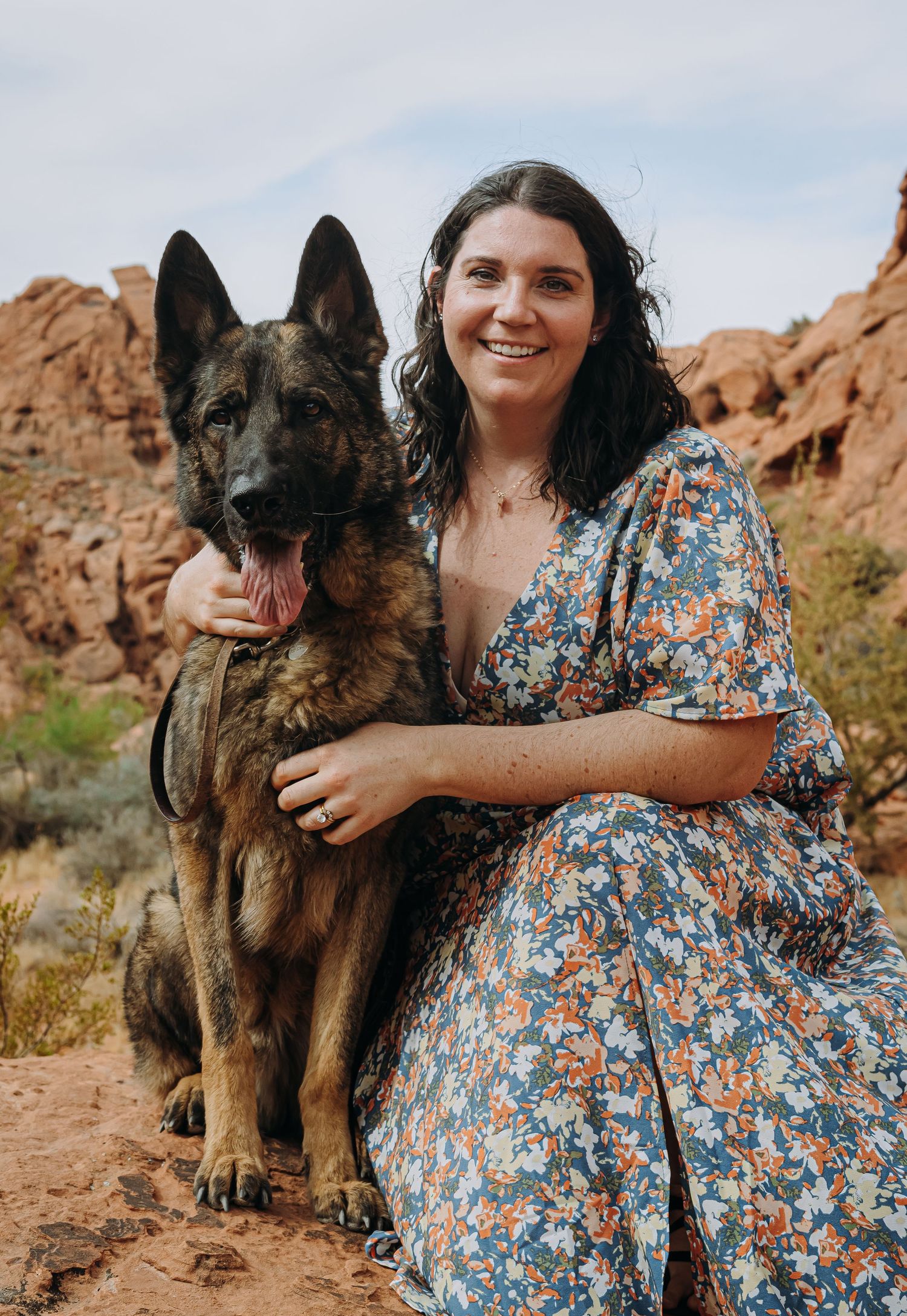 Smiling woman in floral dress posing with her German Shepherd dog in a red rock desert.