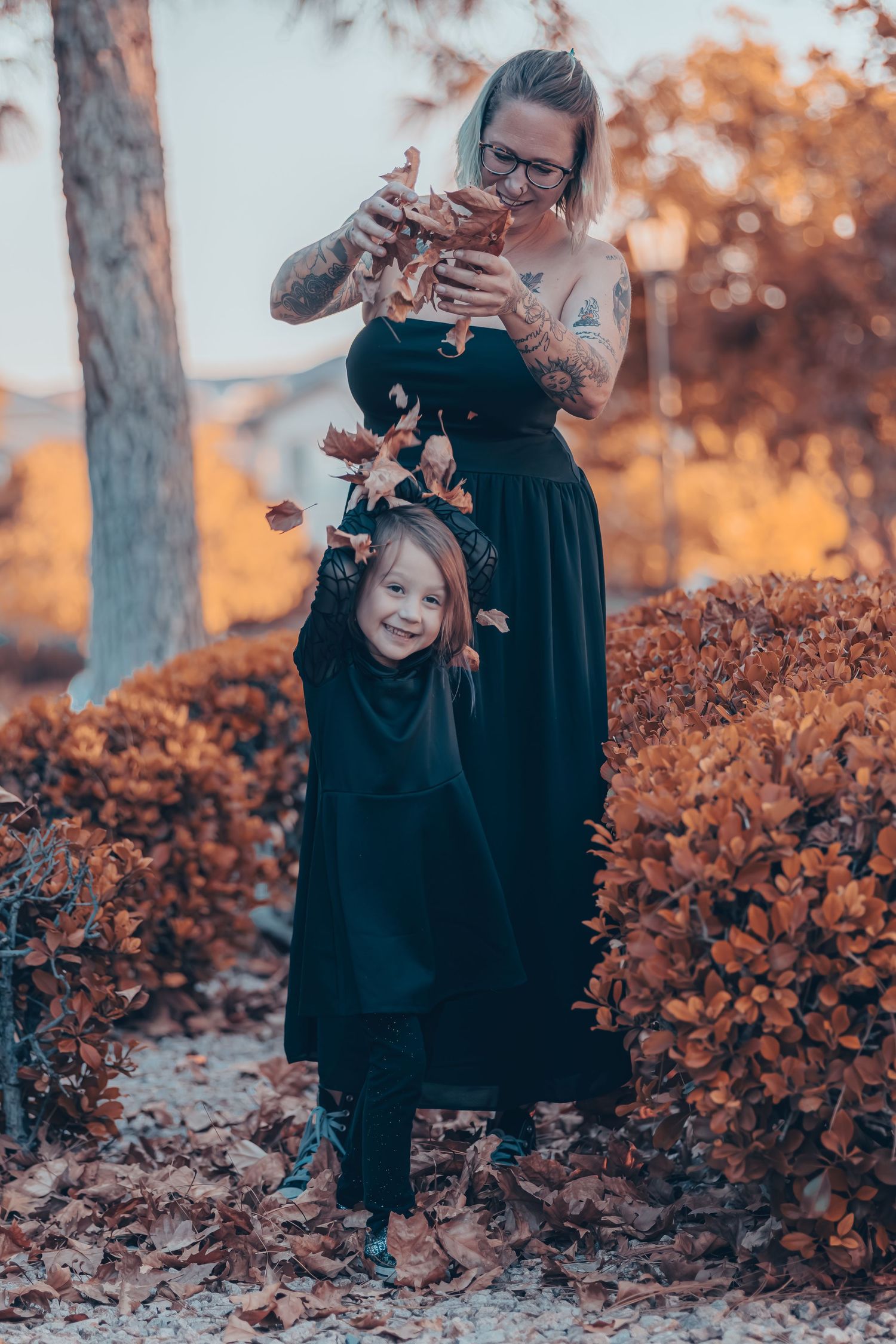 A tattooed mother in black drops autumn leaves over her smiling little girl in a black costume outdoors.