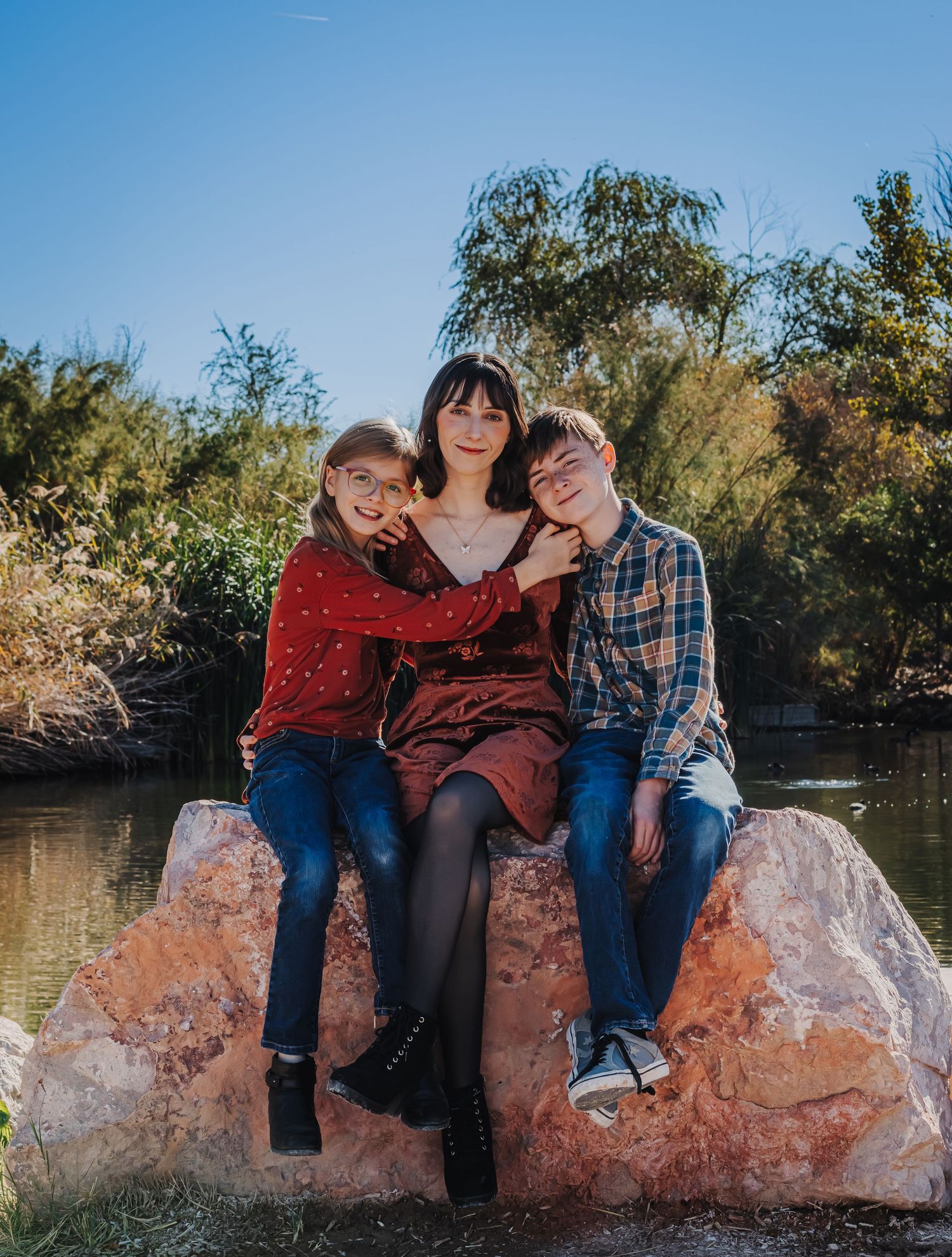 Mother sitting on a rock with two children by a peaceful pond in nature.