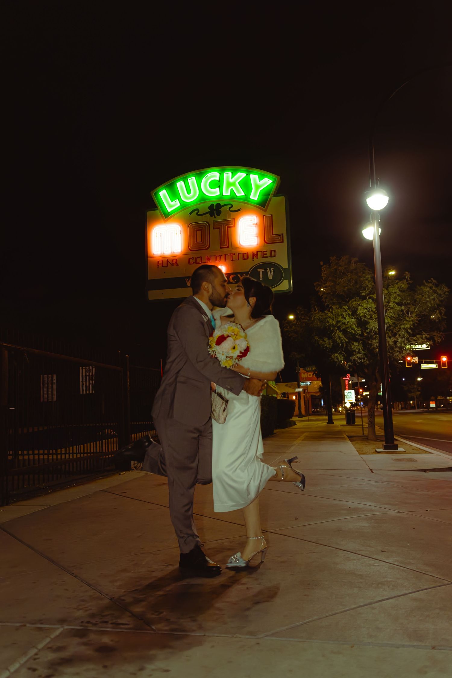 Newlyweds share a romantic kiss under the glowing Lucky neon sign on a Las Vegas street at night.