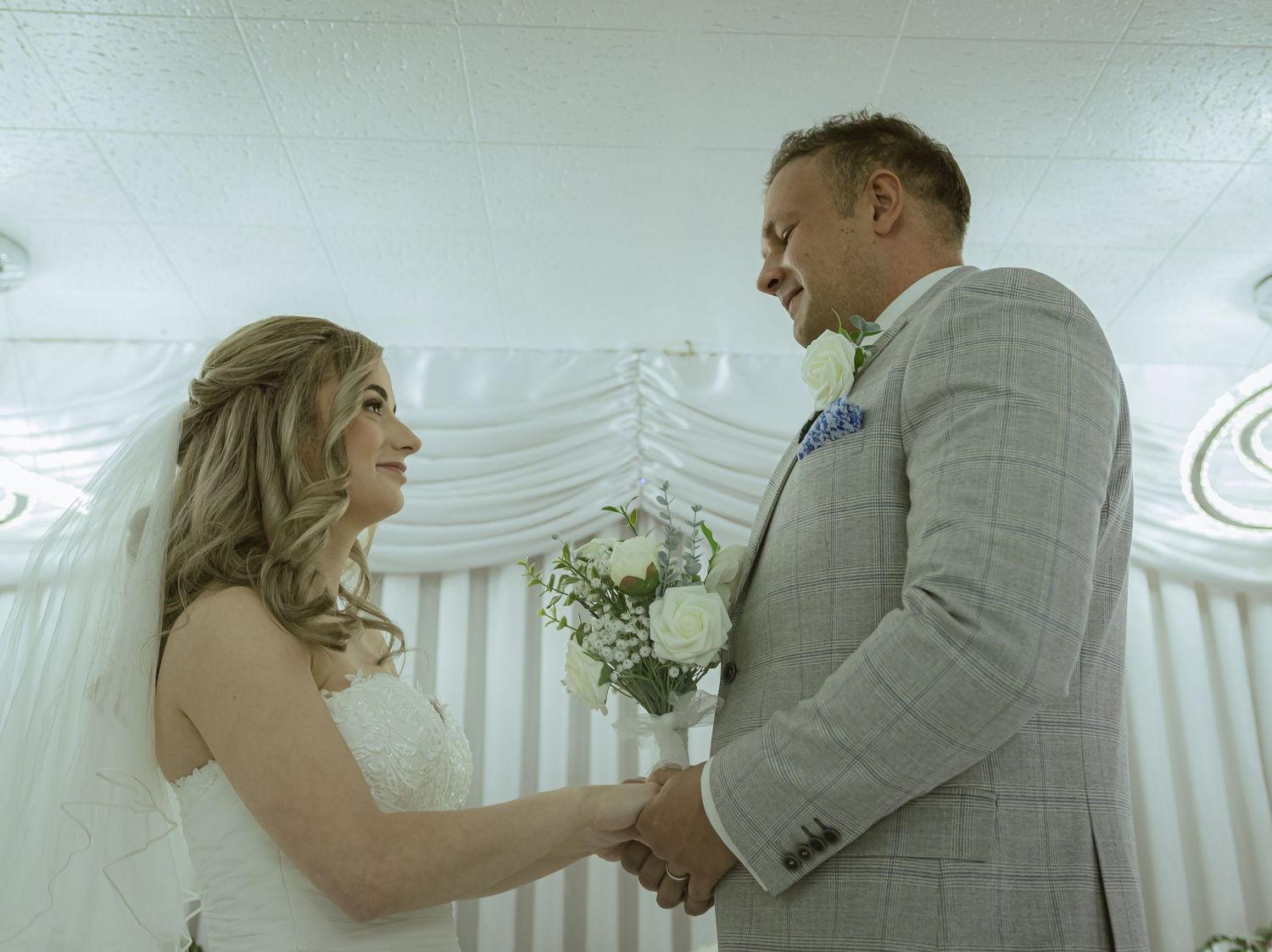 Bride and groom holding hands and smiling at each other during intimate indoor wedding ceremony.