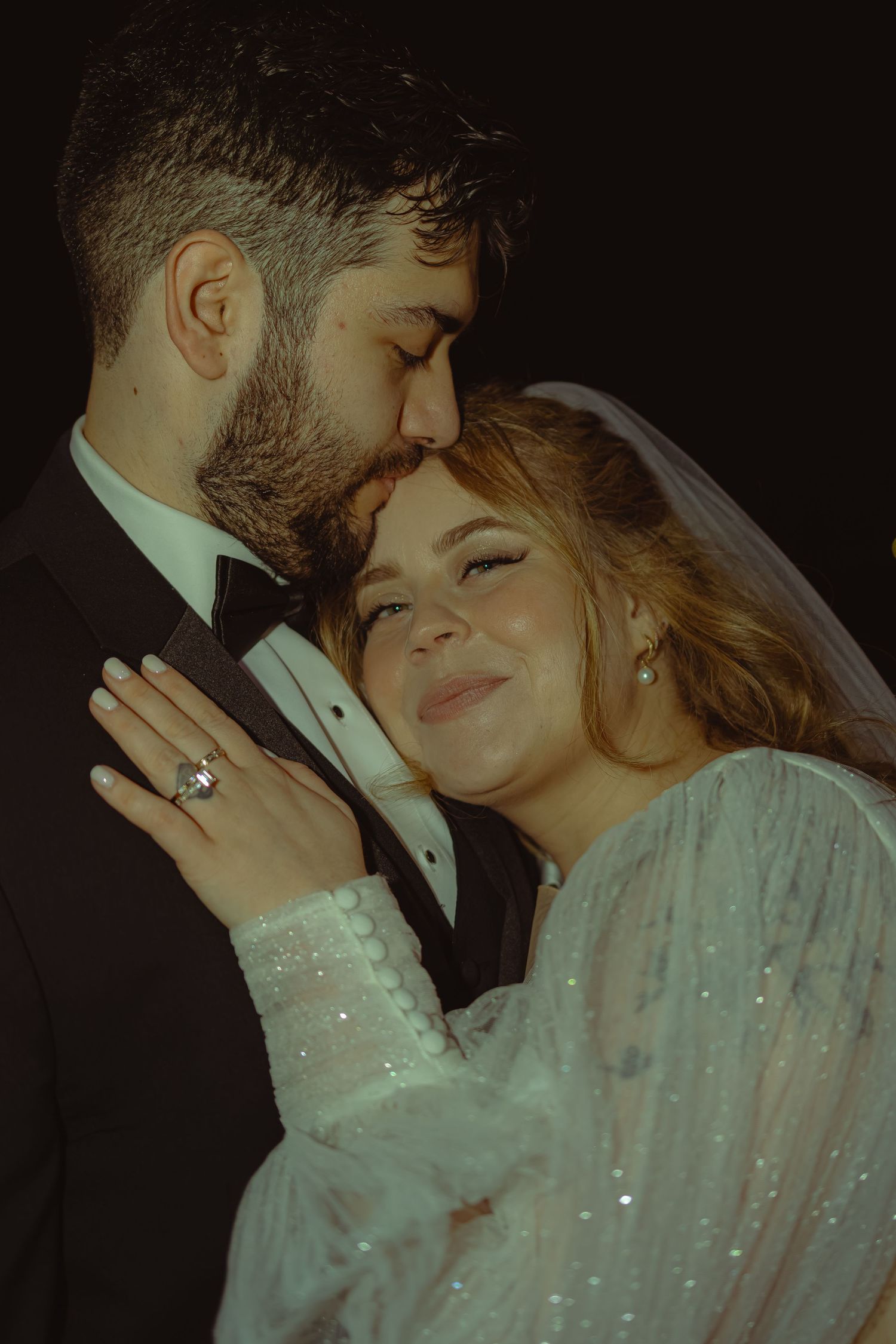 Romantic wedding couple portrait with groom kissing bride's forehead, bride smiling in sparkly white gown with veil.