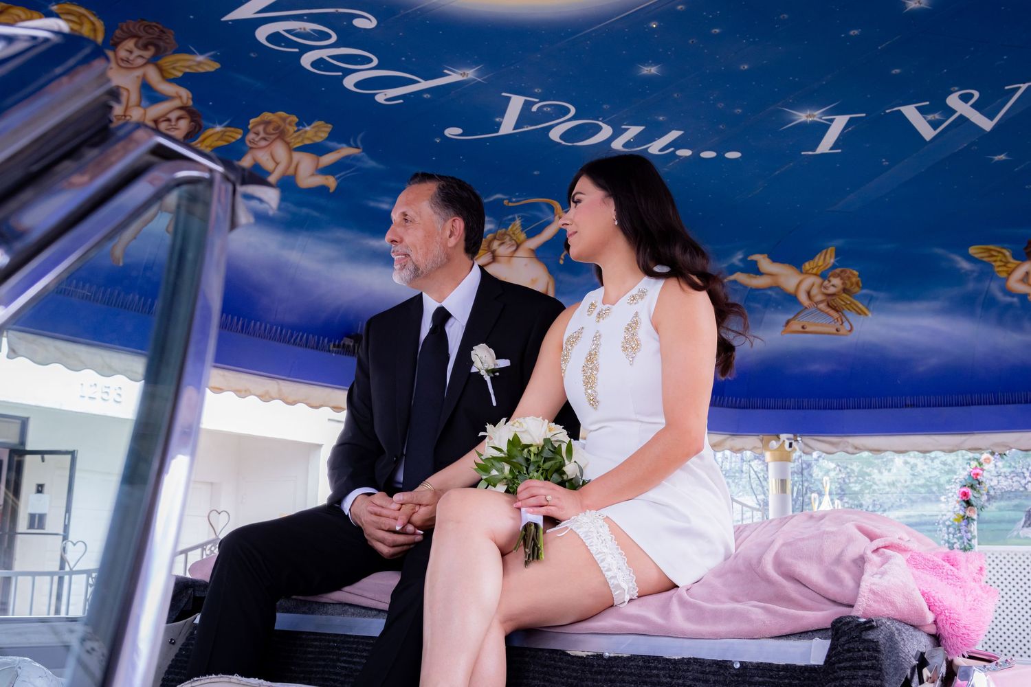 Newlyweds sitting together on a pink car under a cherub-painted ceiling at a Las Vegas chapel.