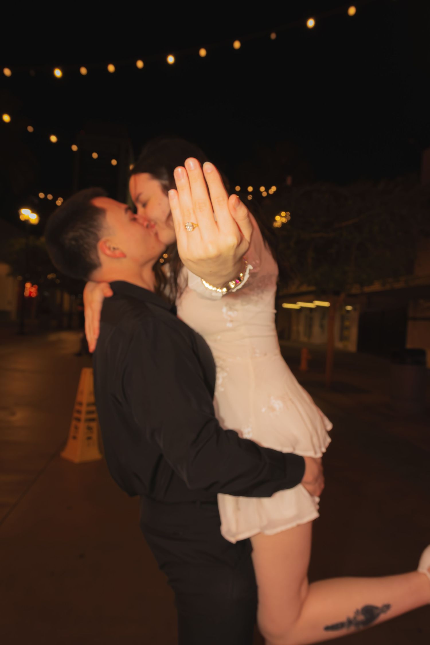 Couple kissing while woman shows off engagement ring under string lights at night in Las Vegas.
