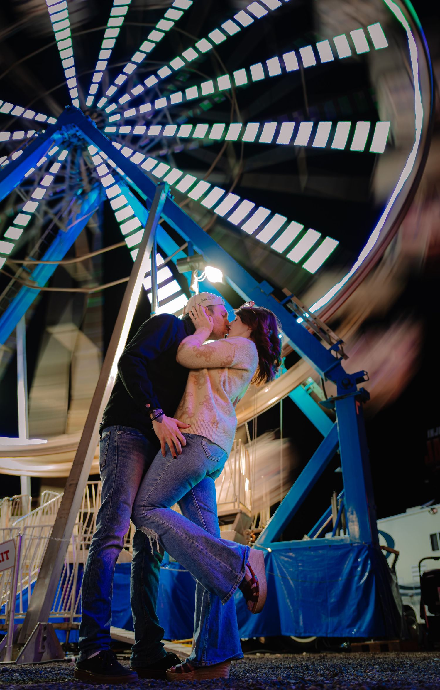 Couple kissing romantically at night in front of a spinning illuminated Ferris wheel at a carnival.