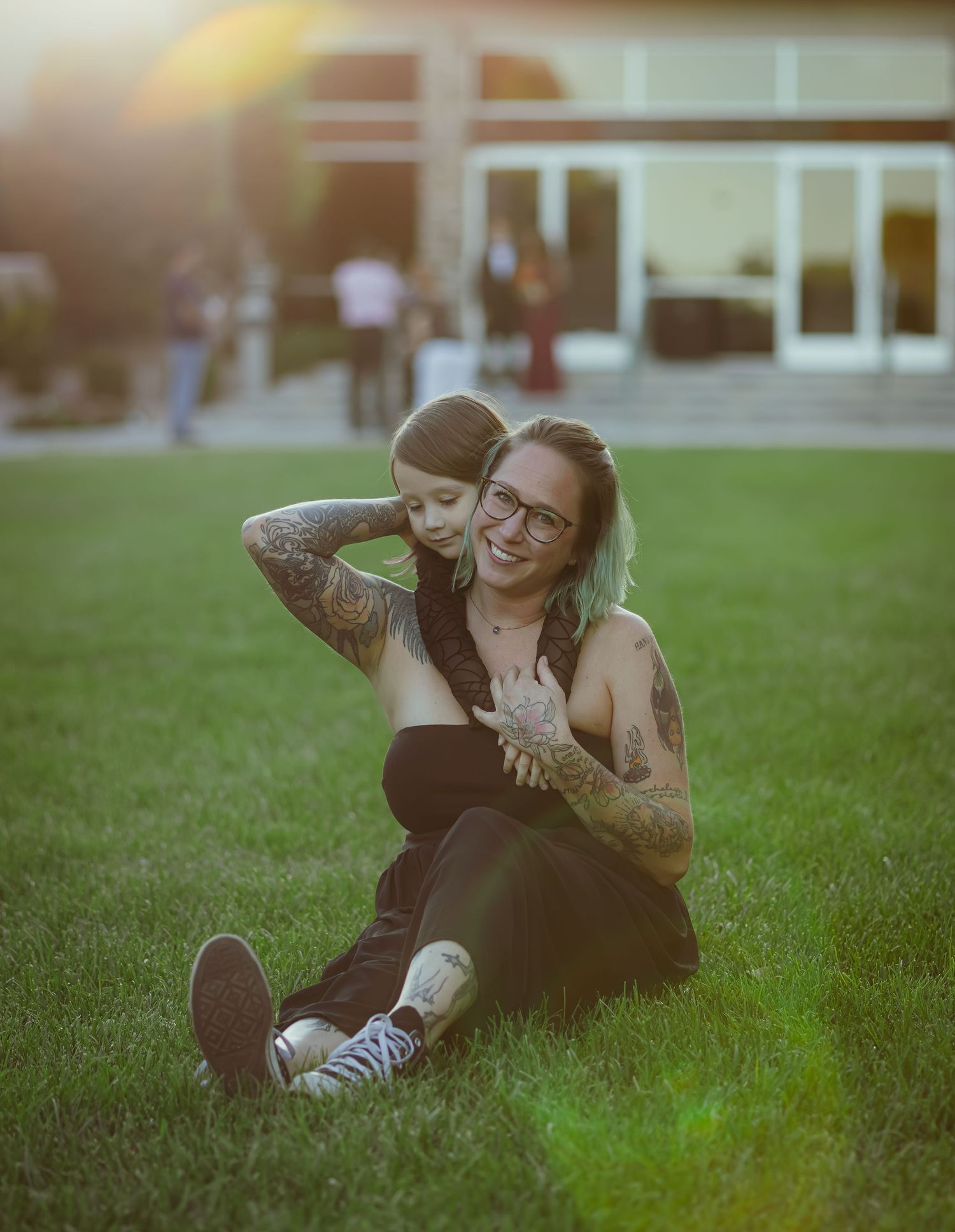 A tattooed woman sits on green grass laughing as a young child hugs her from behind at golden hour.