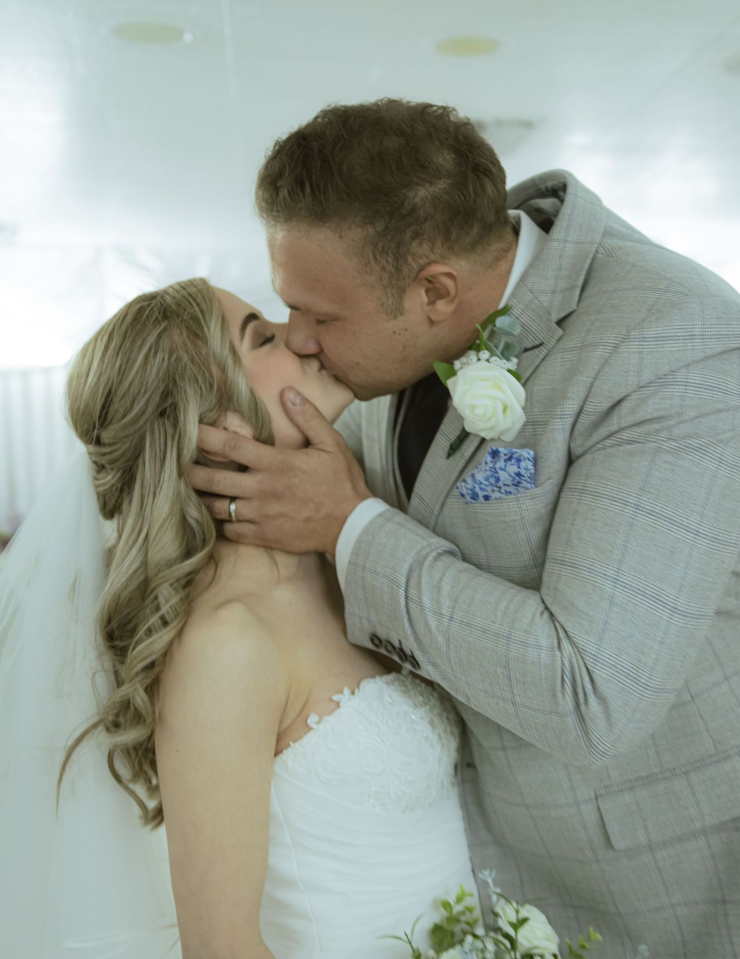 Bride and groom sharing first kiss, groom in grey suit with white boutonniere, bride in white gown.