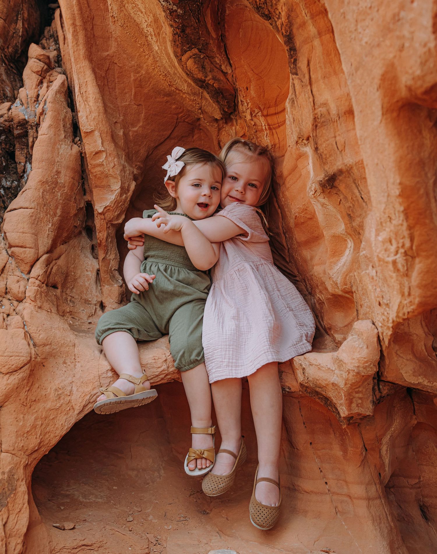 Two young siblings hugging each other while sitting together in a red rock crevice.