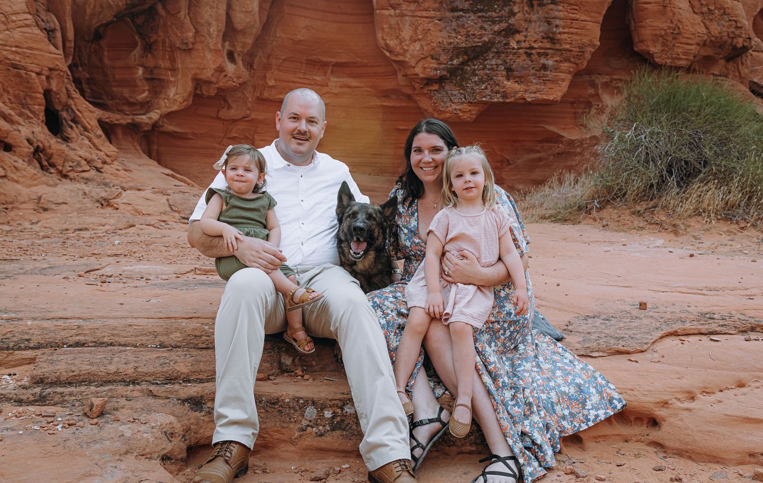 Family with two children and a German Shepherd dog sitting on red sandstone rocks in a desert canyon.