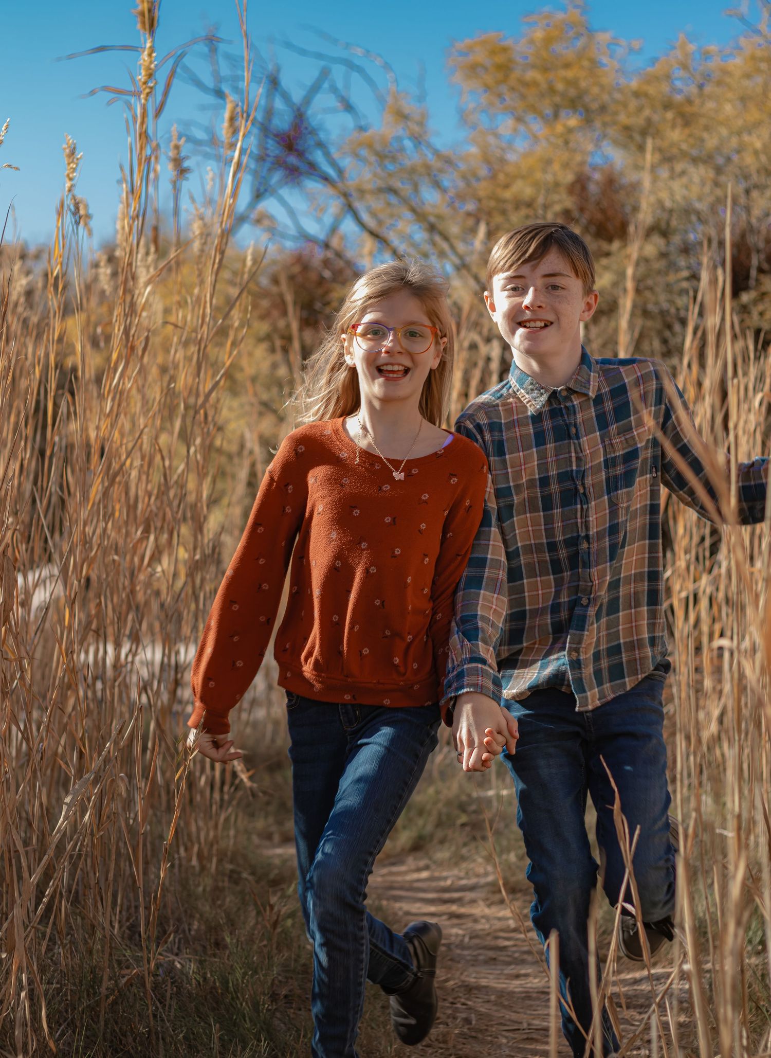 Two happy children run hand-in-hand along a nature trail surrounded by tall golden reeds on a sunny autumn day.