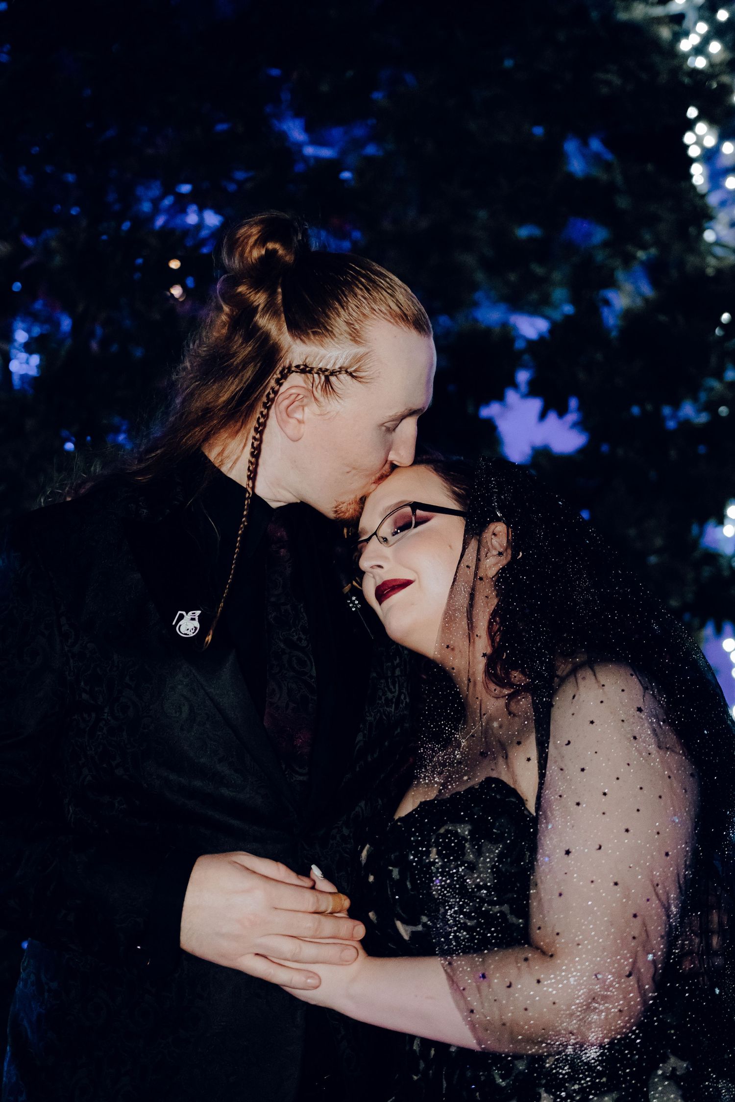Same-sex couple sharing tender kiss at night, both wearing elegant black wedding attire with sparkle details.