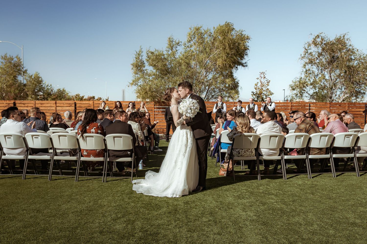 Bride and groom embrace on lawn surrounded by guests seated at long tables at outdoor reception.