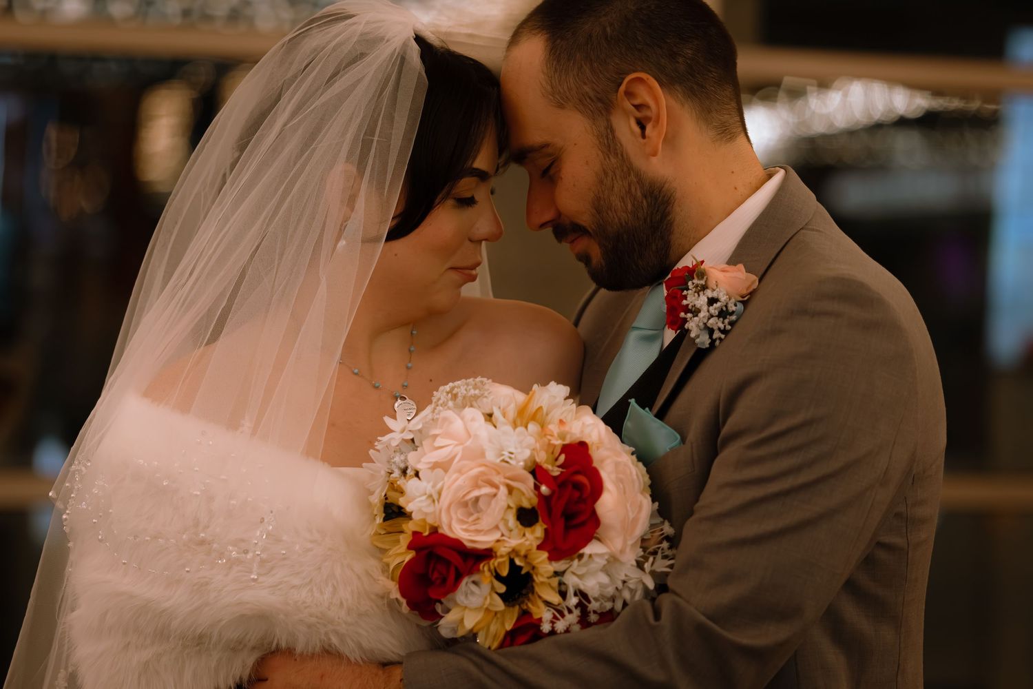 Bride and groom touch foreheads tenderly, bride holding a lush bouquet of red and white roses.