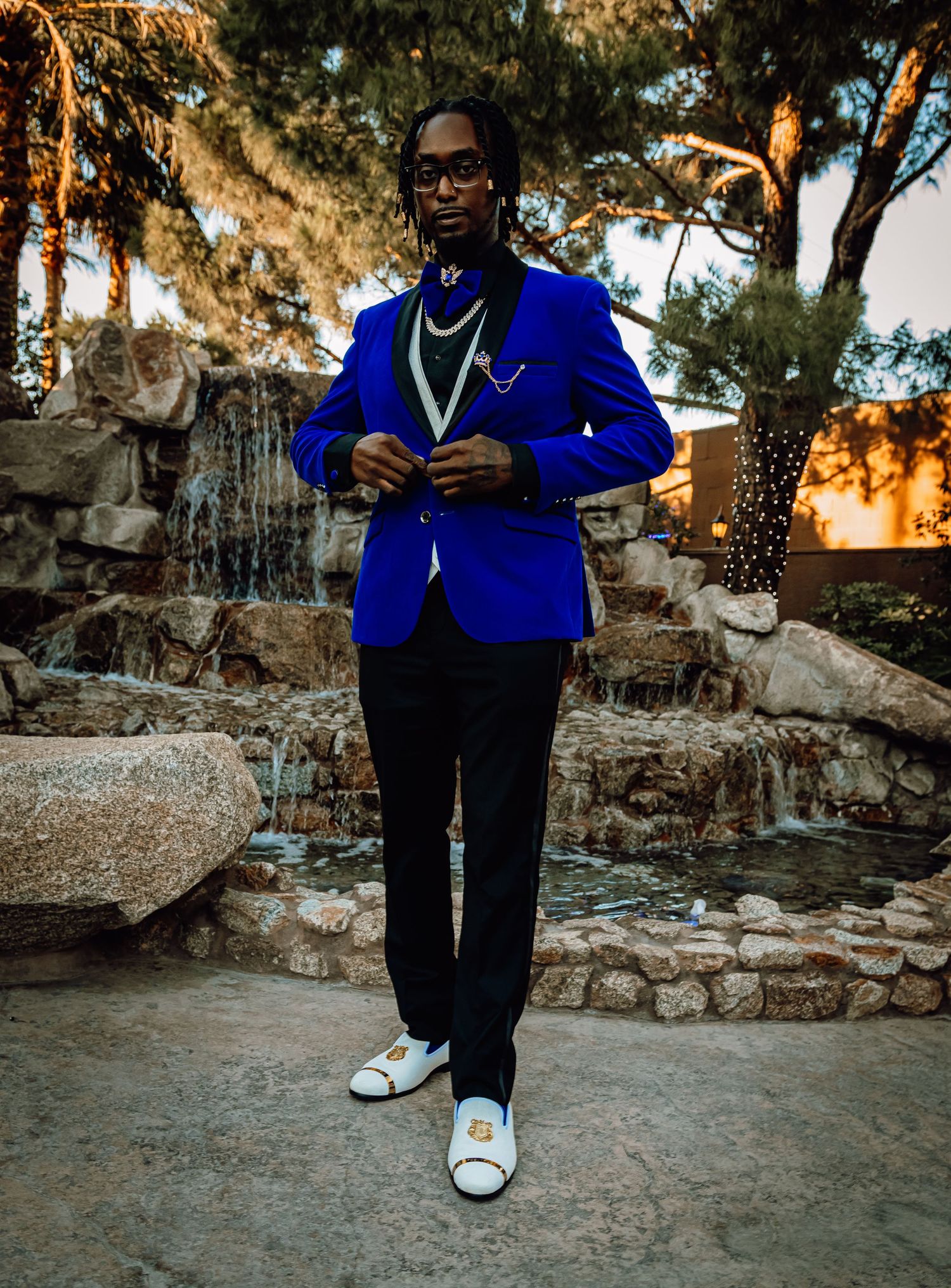 Man in royal blue velvet blazer with pearl necklace posing by stone fountain at golden hour.