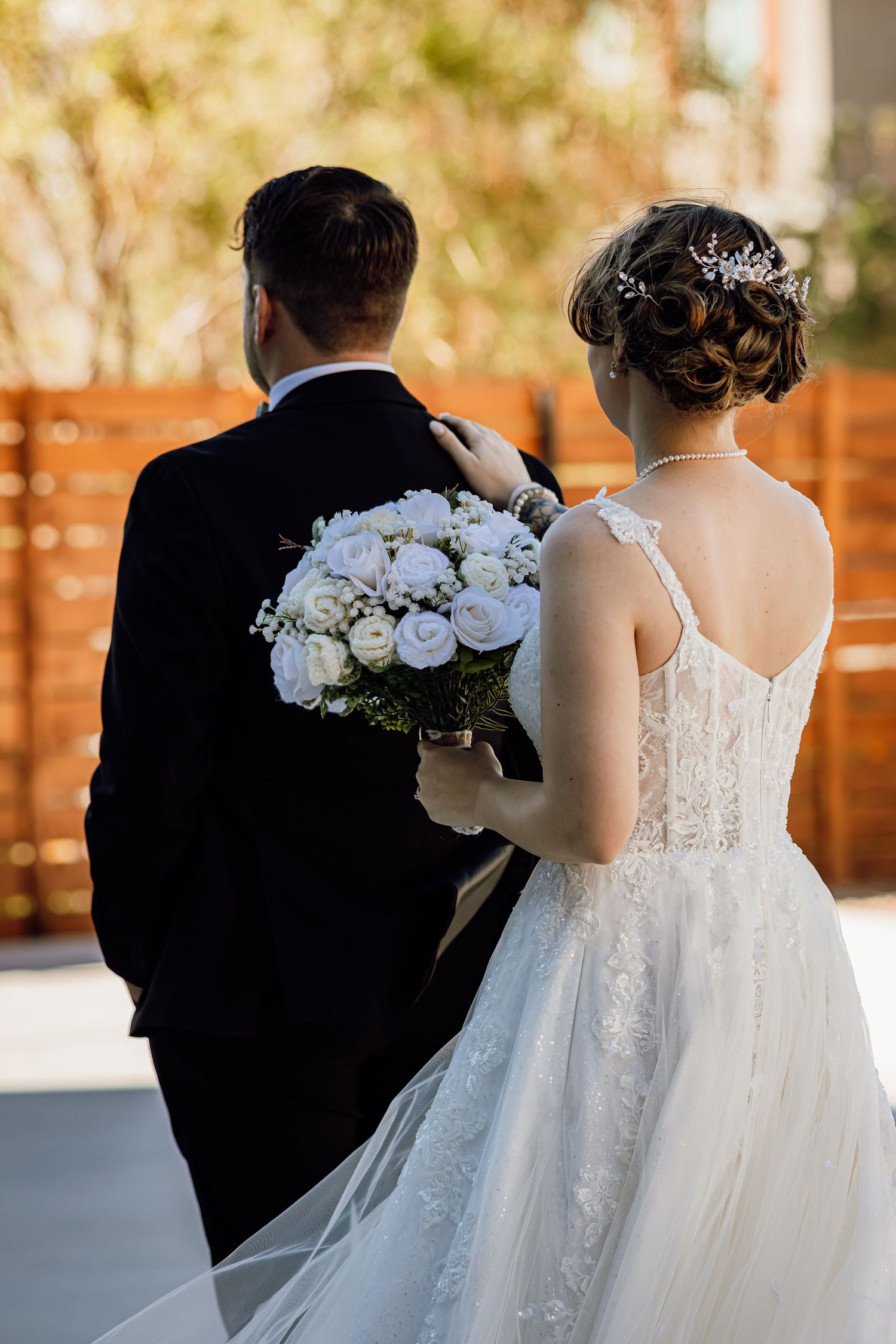 Bride in lace gown and groom walk away together, bride carrying a white rose bouquet outdoors.