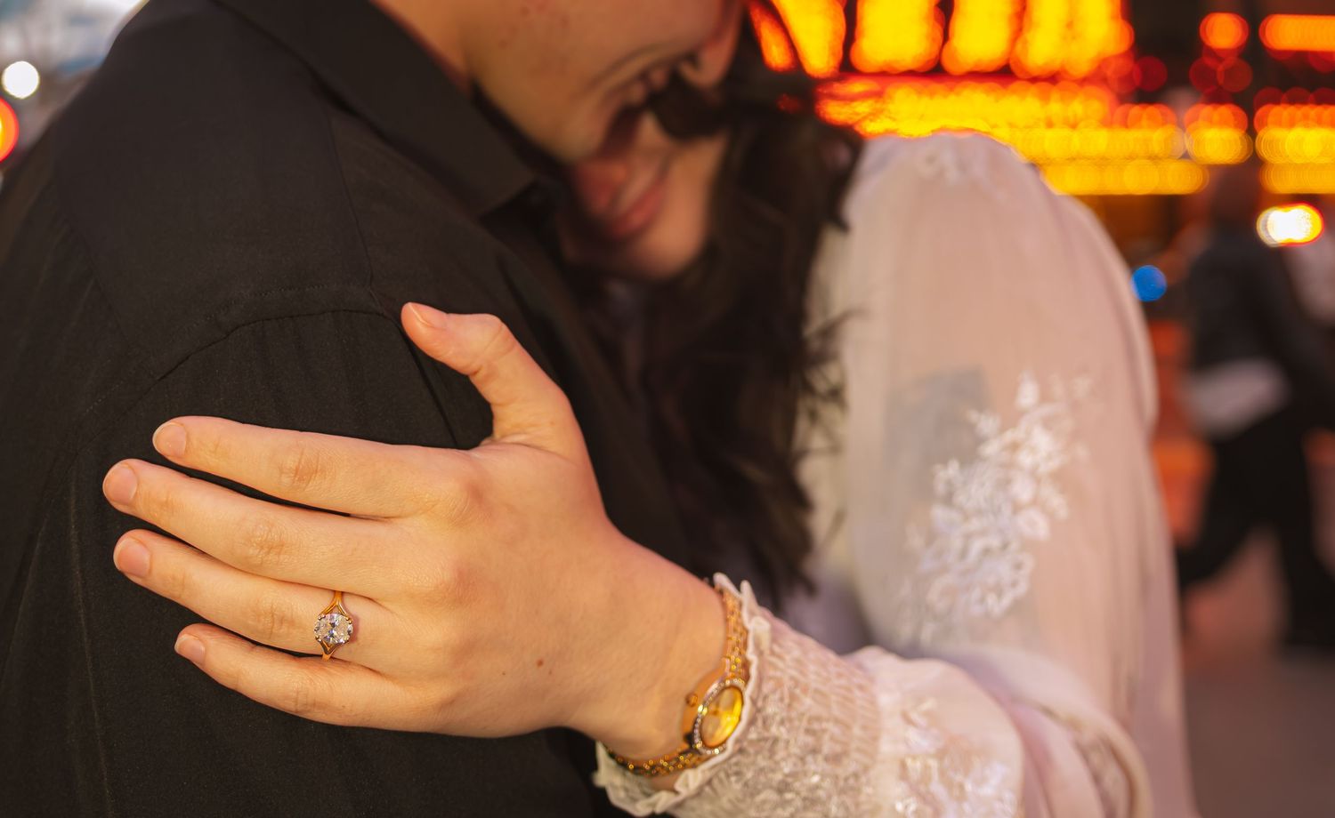 Close-up of a couple embracing showing an engagement ring and gold bracelet against neon-lit Las Vegas backdrop.